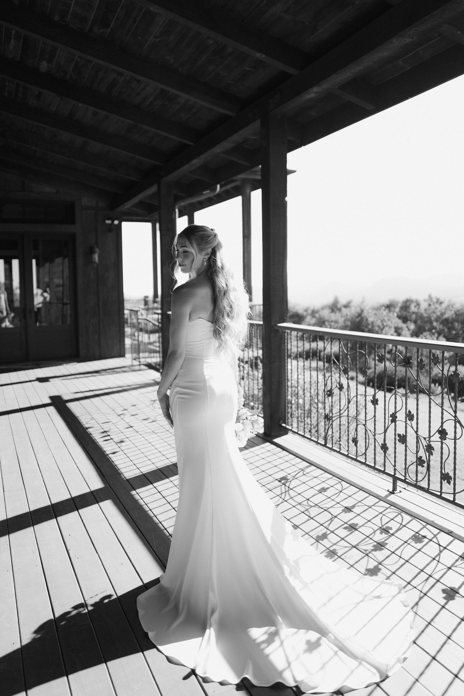 Elegant bride in fitted white gown standing on rustic balcony, black and white portrait.