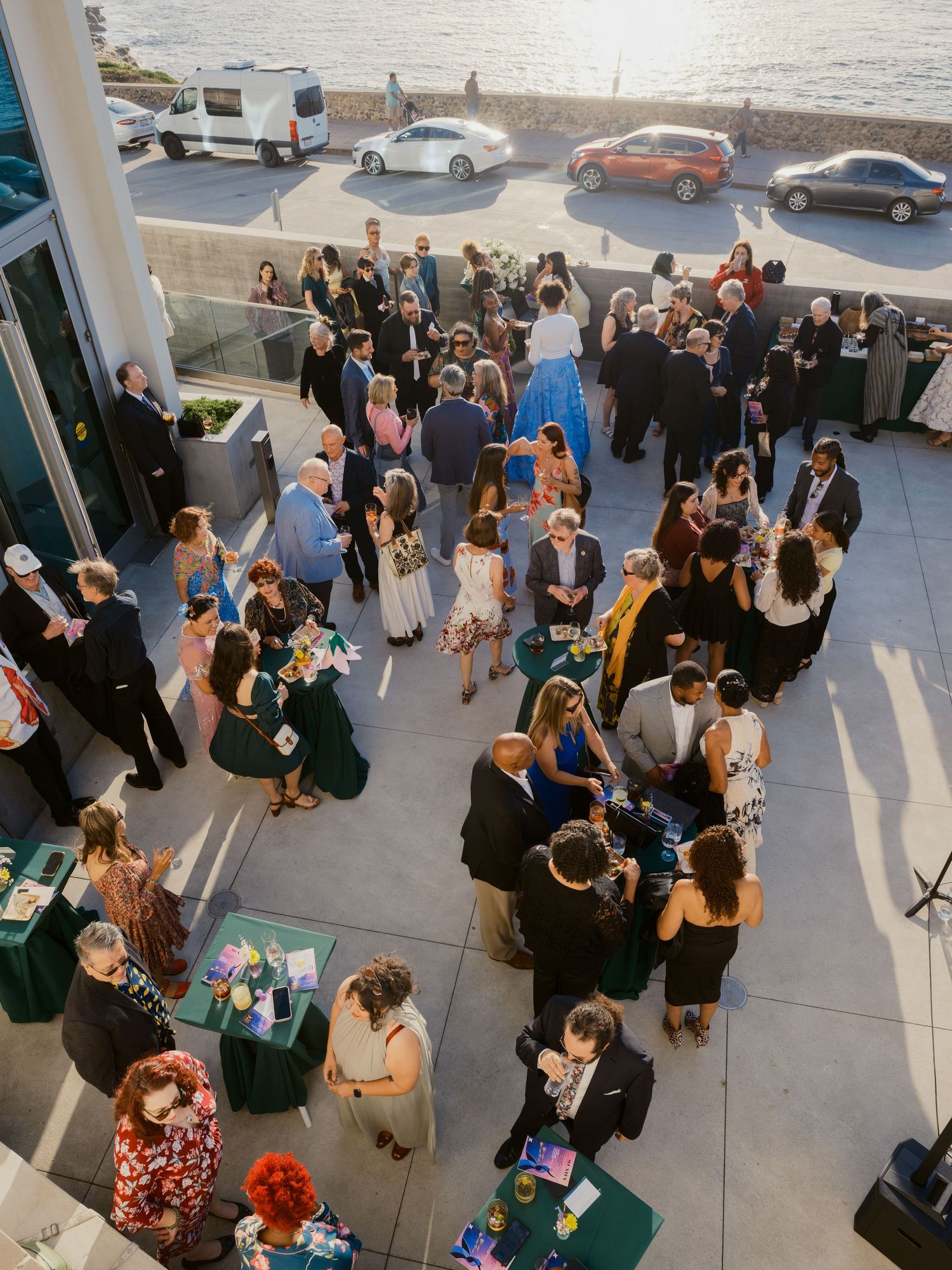 Aerial view of elegantly dressed guests mingling on a waterfront terrace during an outdoor evening social event.