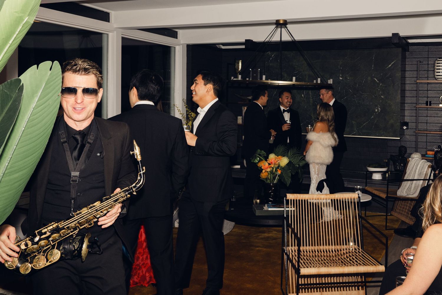 Saxophonist in black suit performs at elegant wedding reception with guests mingling in stylish venue.