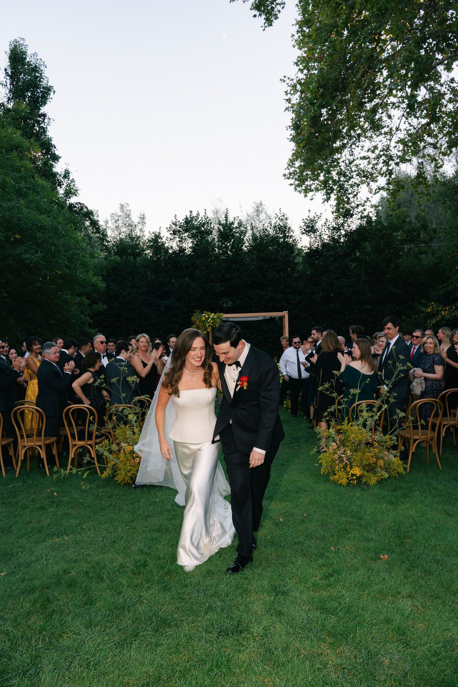 Smiling bride in a white gown and groom in a black tuxedo walk down the grass aisle past applauding guests at an outdoor garden wedding.