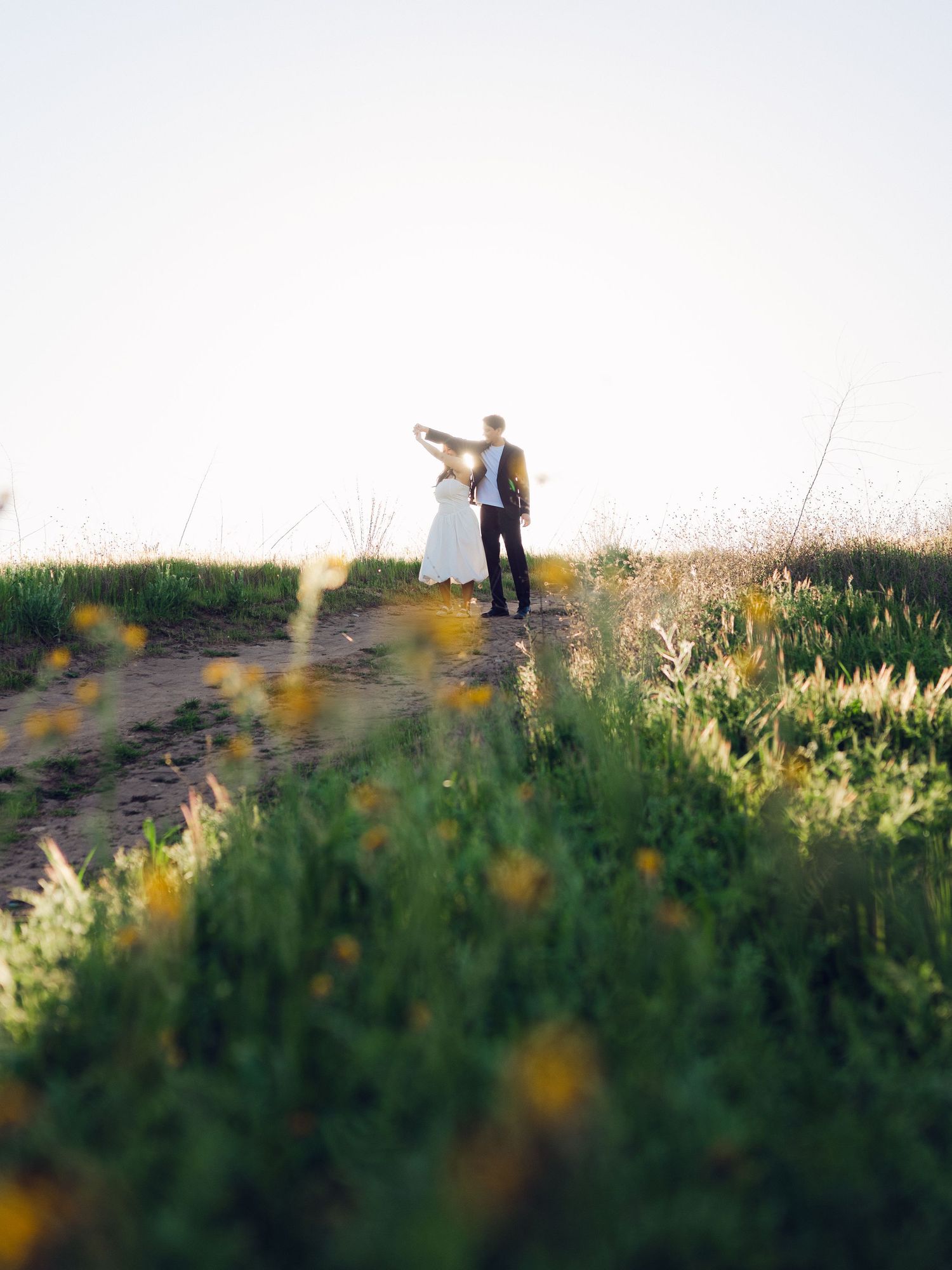 Bride and groom dancing together on a scenic dirt path surrounded by wildflowers and golden evening light.