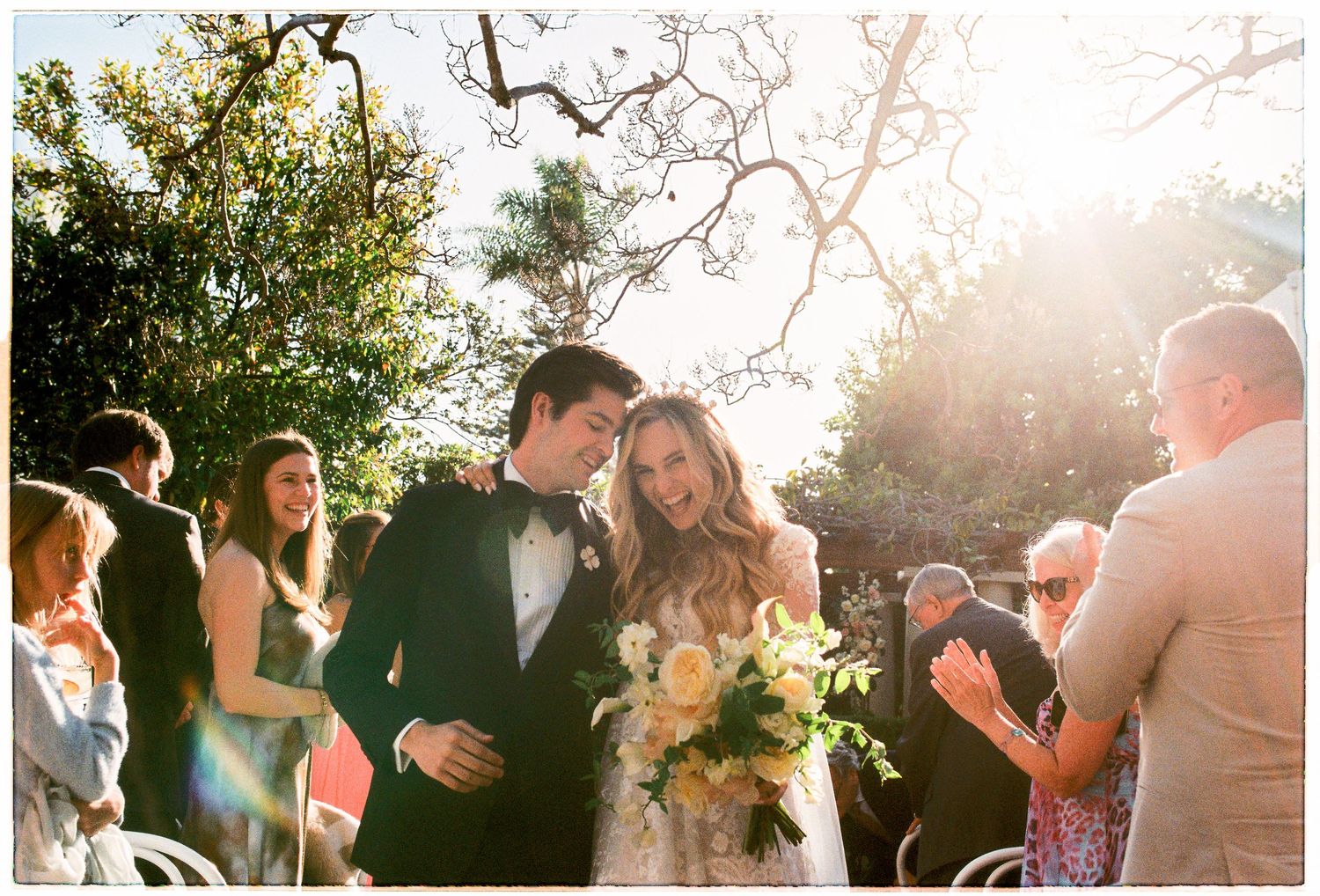 Newlyweds walk back down aisle smiling as guests applaud at sunny outdoor garden wedding ceremony.