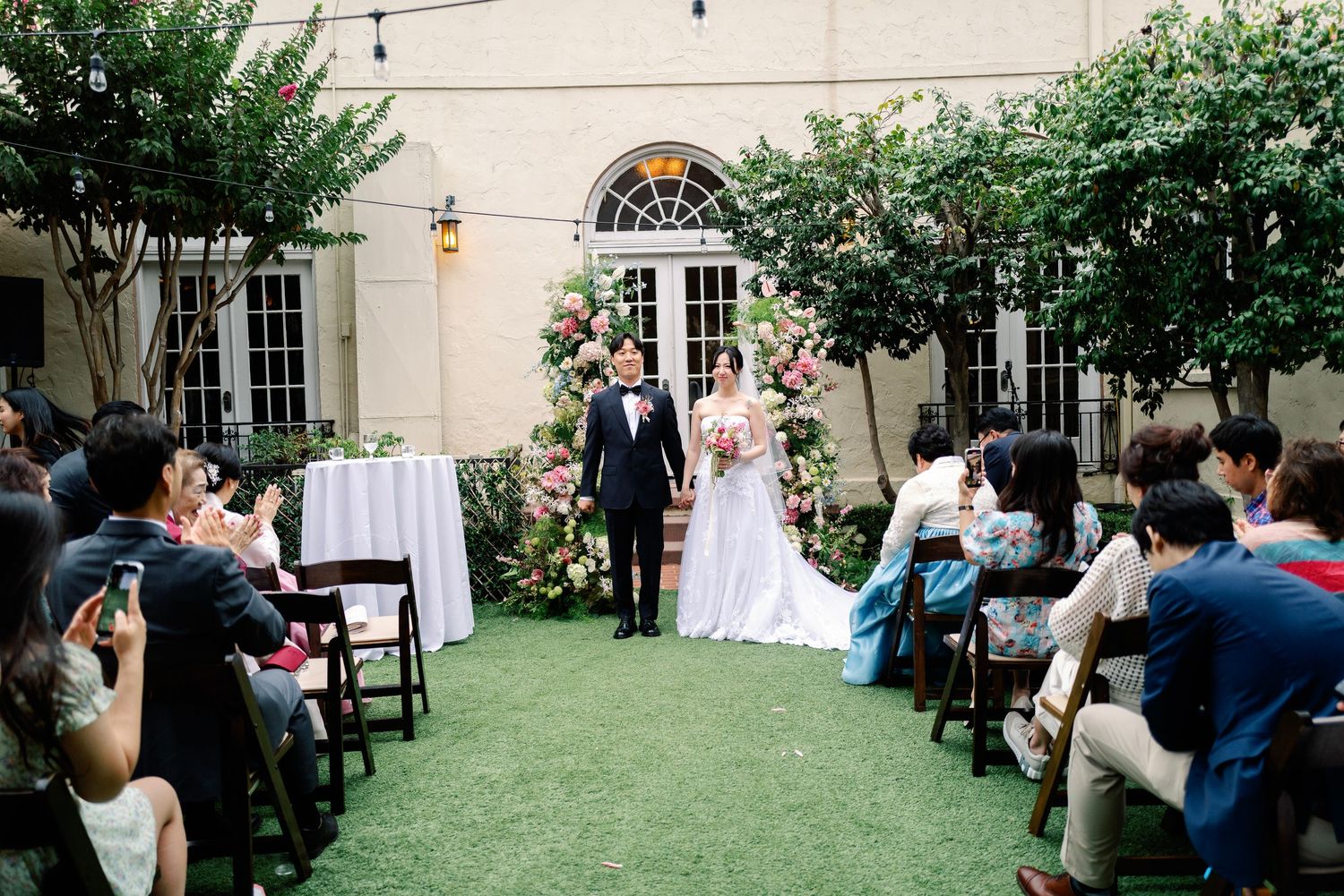 Newlyweds walk down aisle at elegant outdoor garden wedding ceremony with lush floral backdrop.