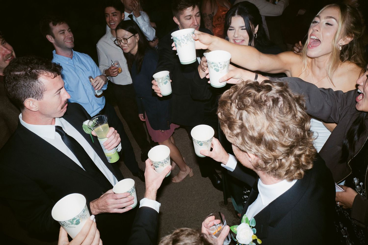 Wedding guests toasting with white cups at a lively indoor reception party.