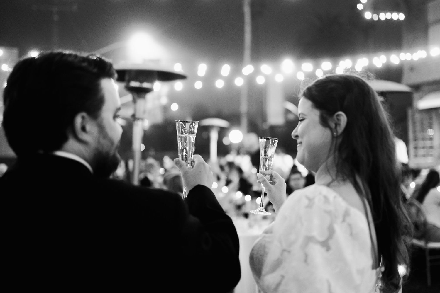 Black and white close-up of bride and groom toasting champagne glasses at outdoor evening wedding reception.