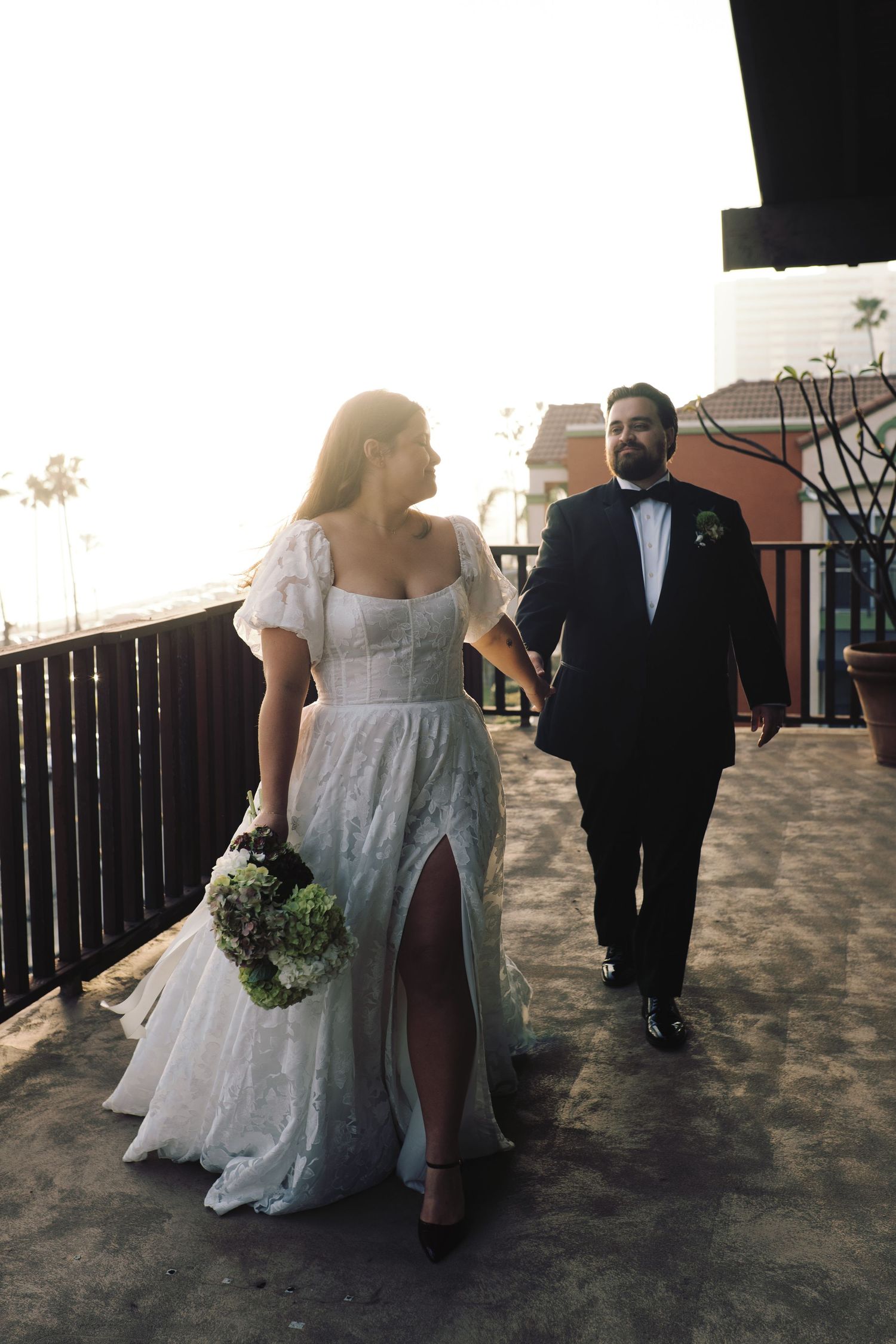Bride and groom walk hand in hand on balcony at golden hour after wedding.