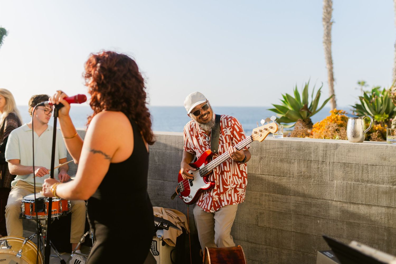 Band performing outdoors near ocean, guitarist in floral shirt, female vocalist with microphone, drummer in background.