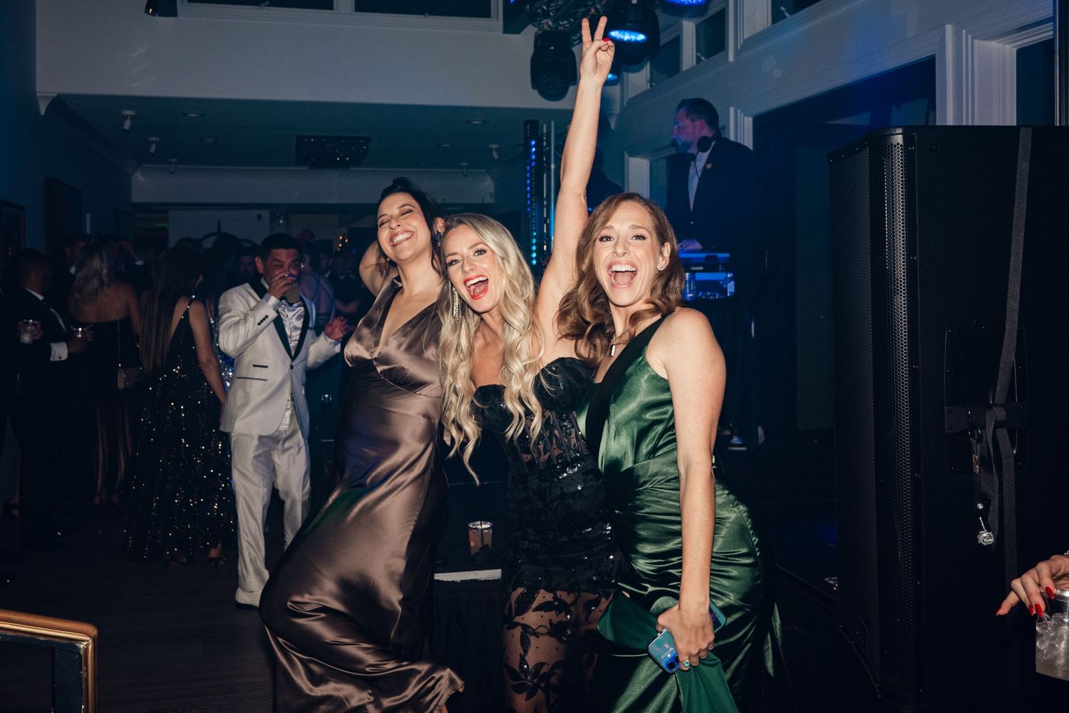Three women in elegant gowns dancing and celebrating at a formal evening party event.