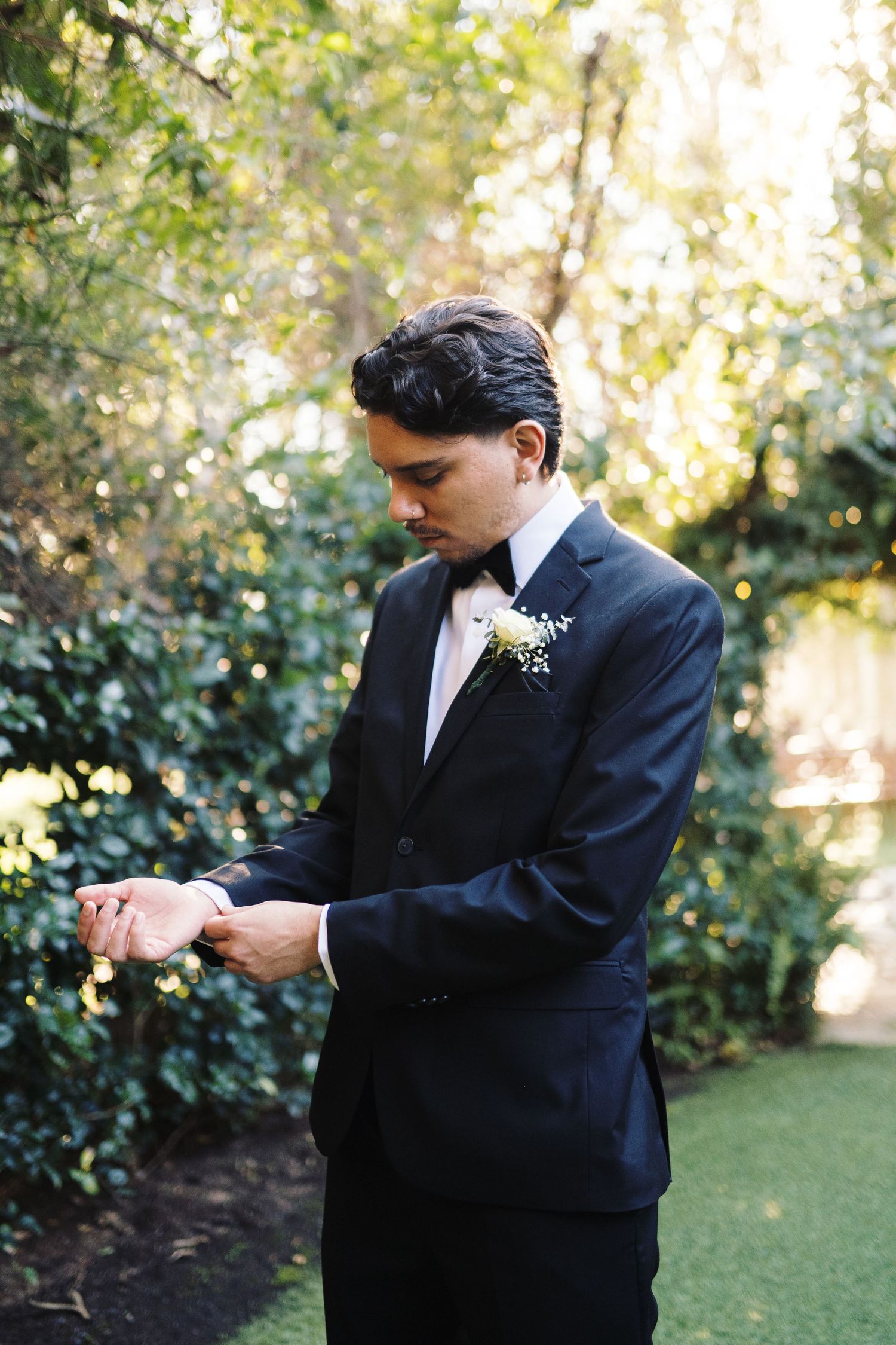 Groom in black tuxedo with floral boutonniere adjusting cufflinks outdoors in lush garden setting.