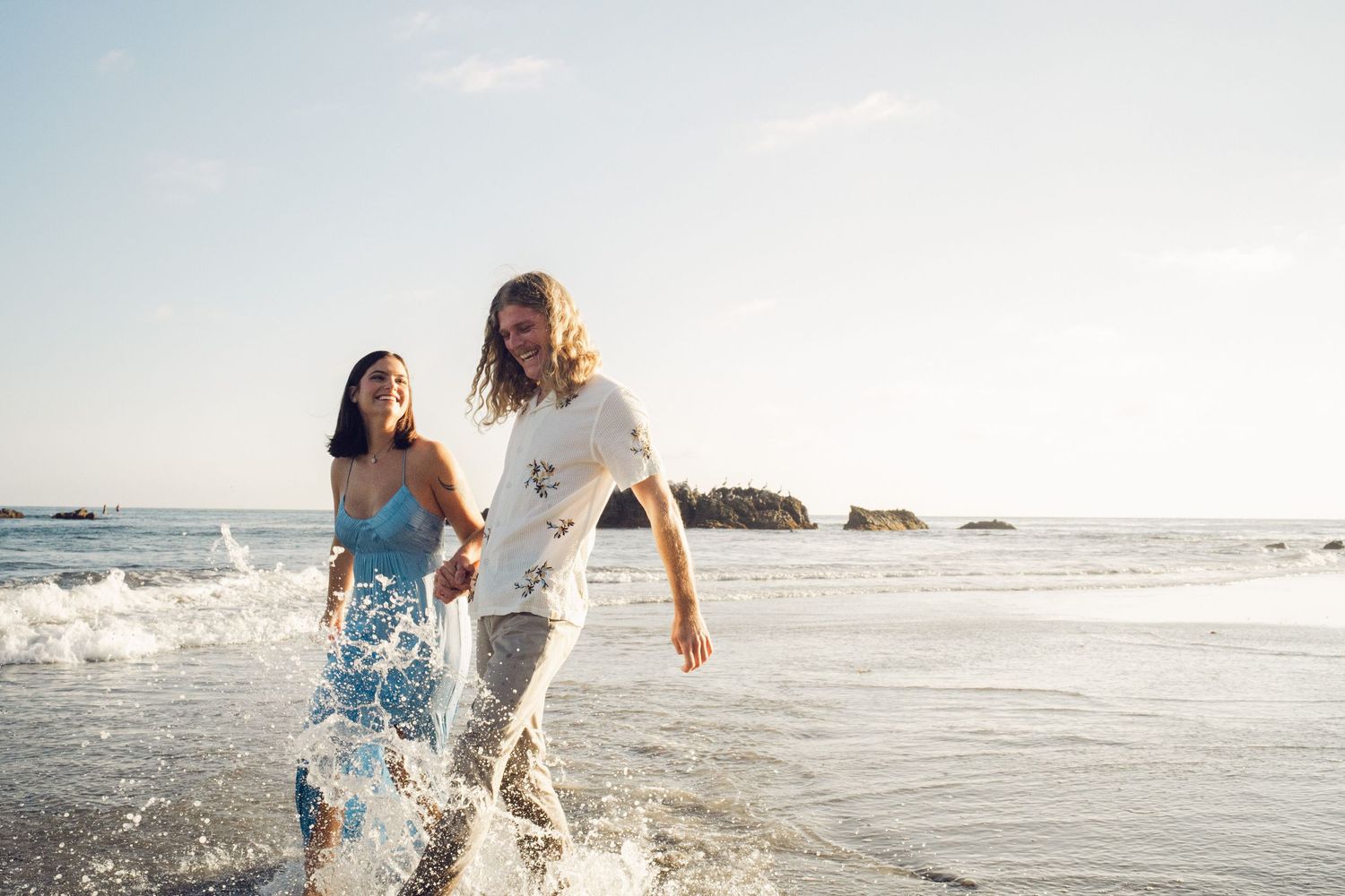 Couple holding hands and splashing through ocean waves on a sunny beach.