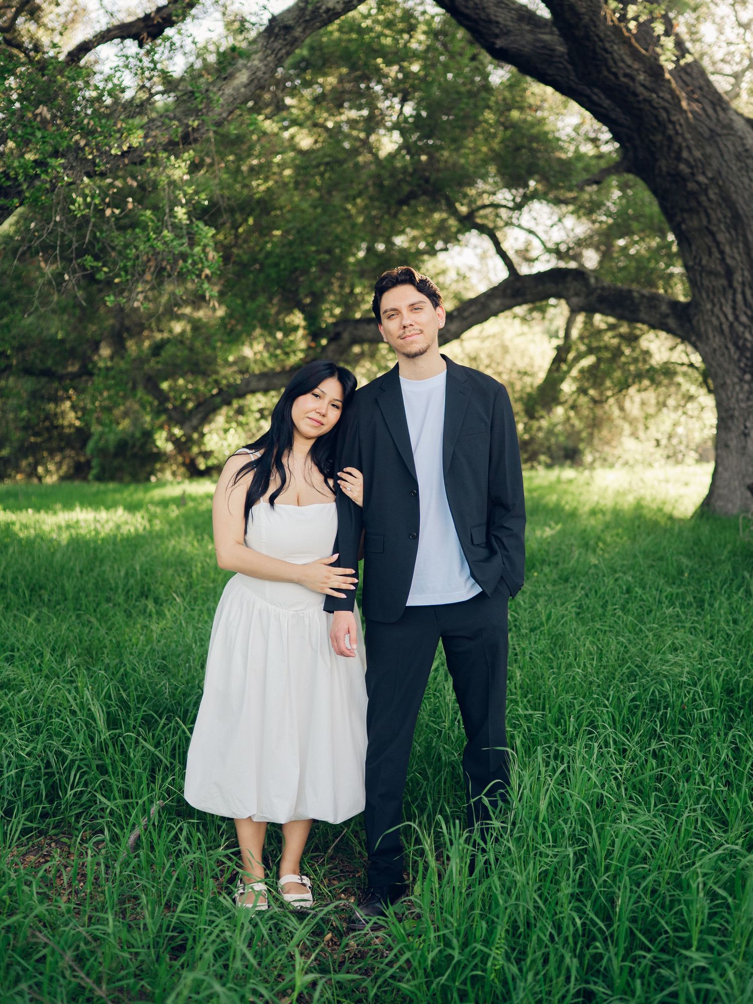 Couple poses together in lush green meadow under oak tree during outdoor engagement session.