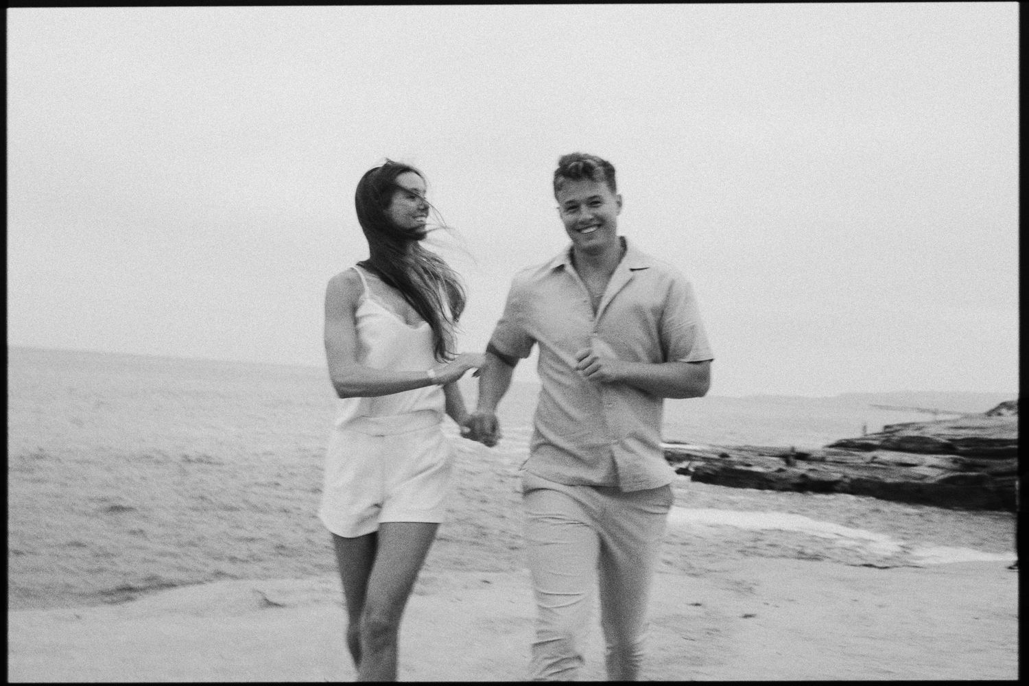 Black and white photo of a smiling couple walking hand in hand along a beach.
