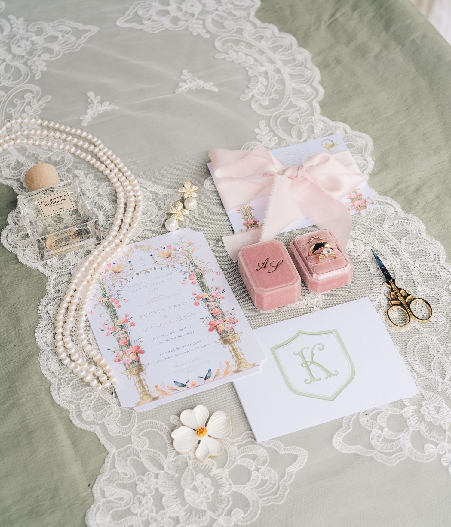 Flat lay of wedding details including floral invitation, pearl necklace, pink velvet ring boxes, perfume, and monogram card on sage linen.