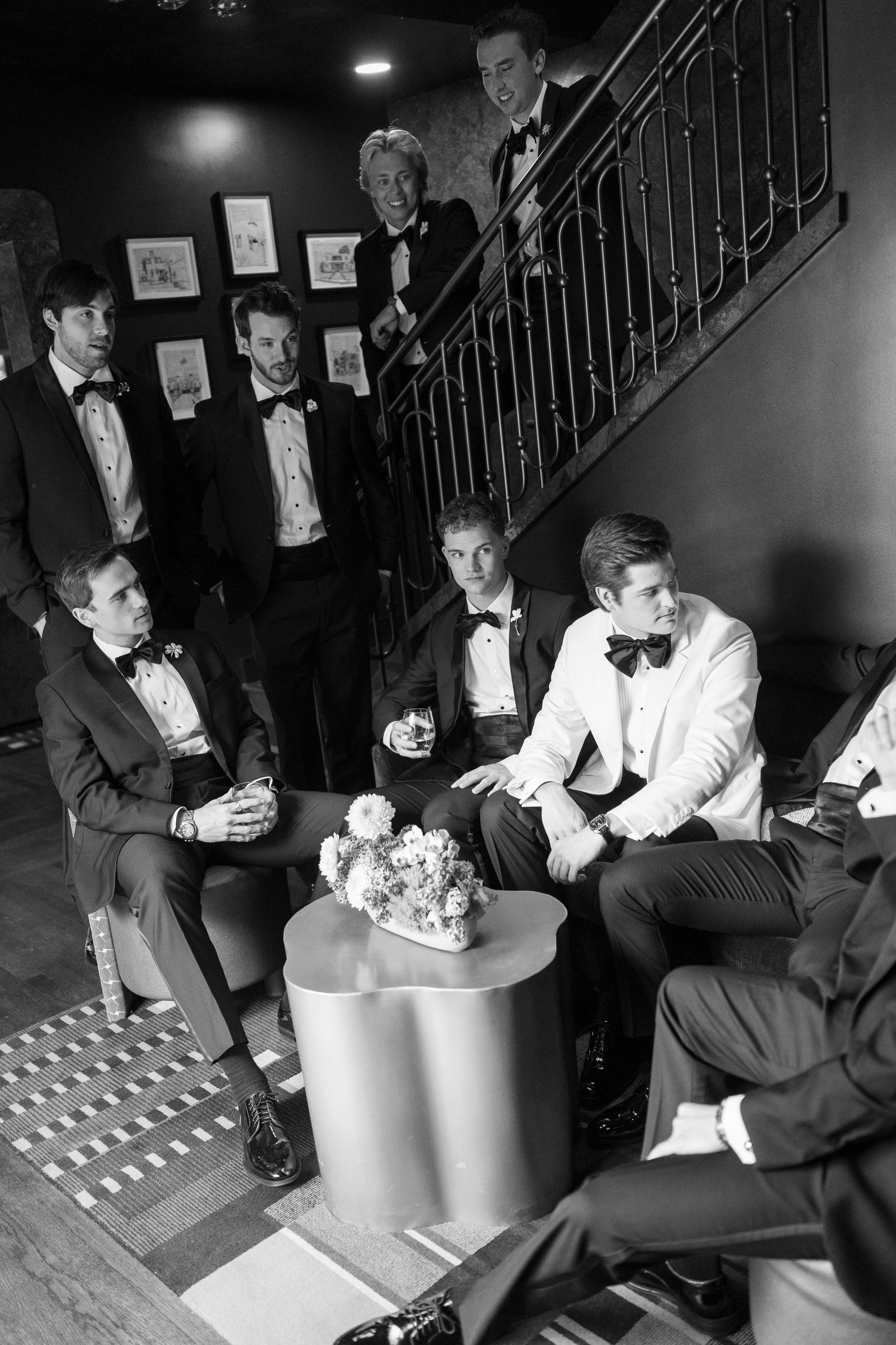 Black and white photo of groomsmen in tuxedos gathered around staircase inside elegant venue before wedding ceremony.