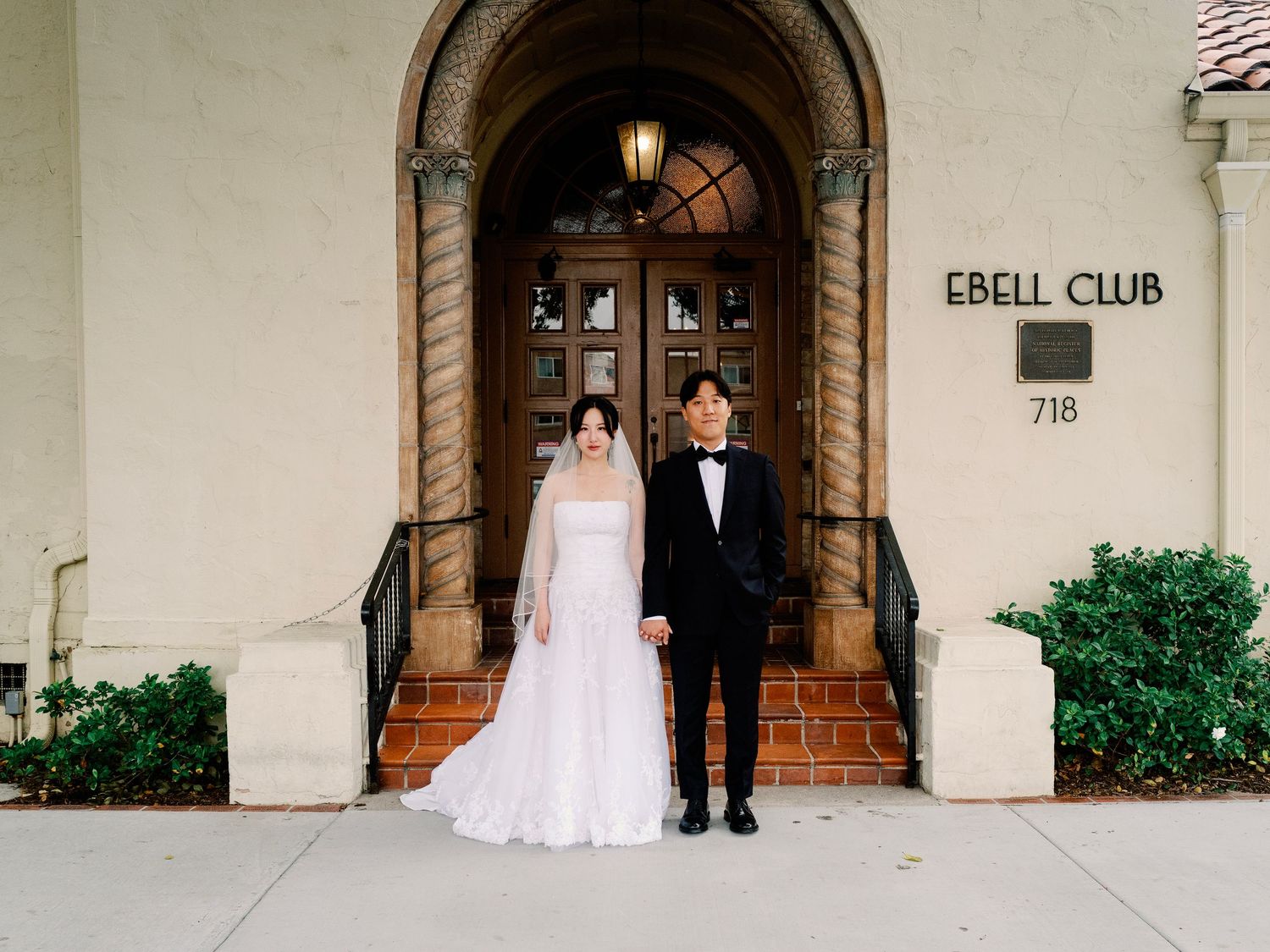 Bride in white gown and groom in tuxedo holding hands at Ebell Club entrance.