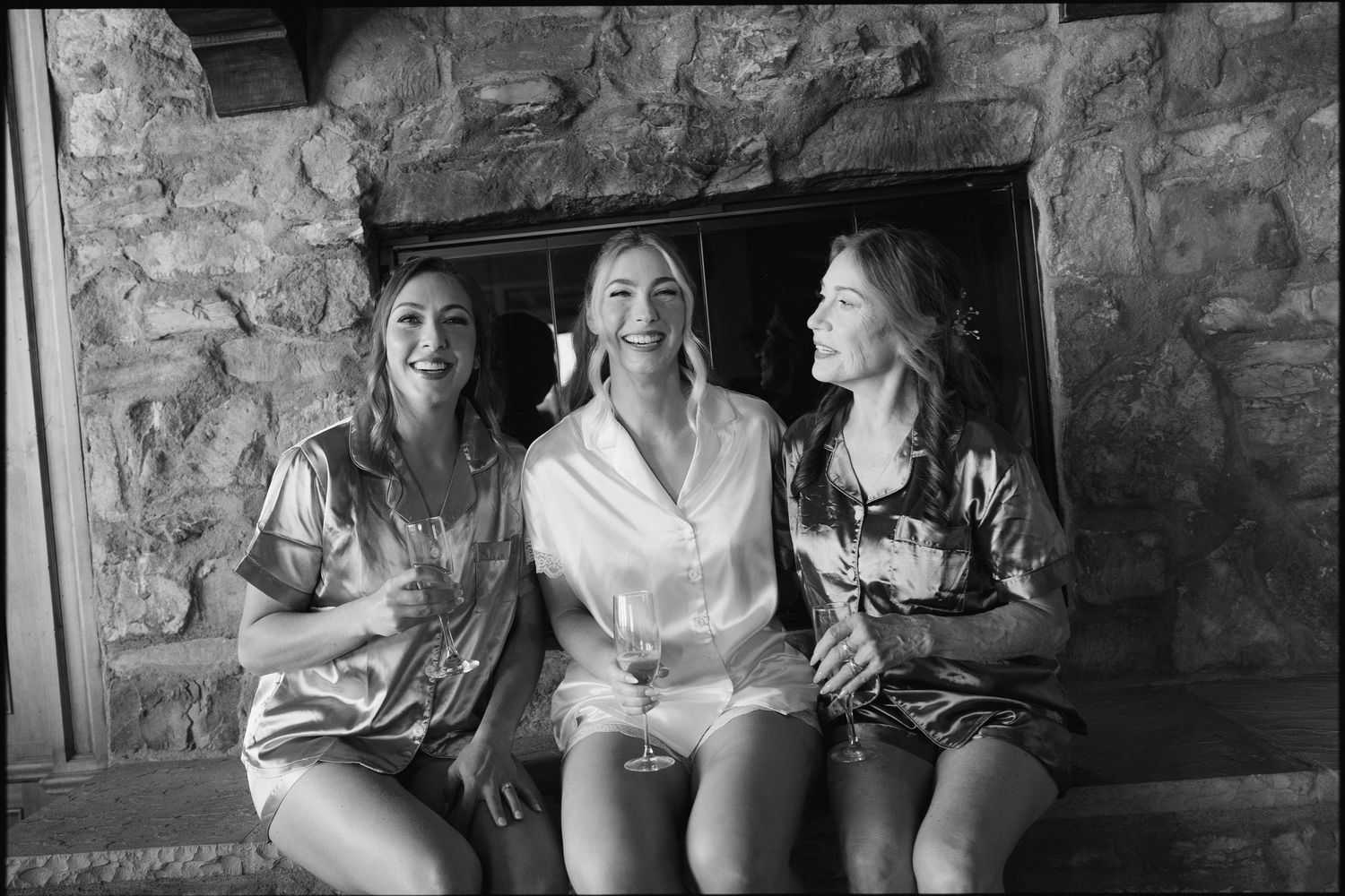 Three bridesmaids laughing together in satin robes by a stone fireplace, black and white.