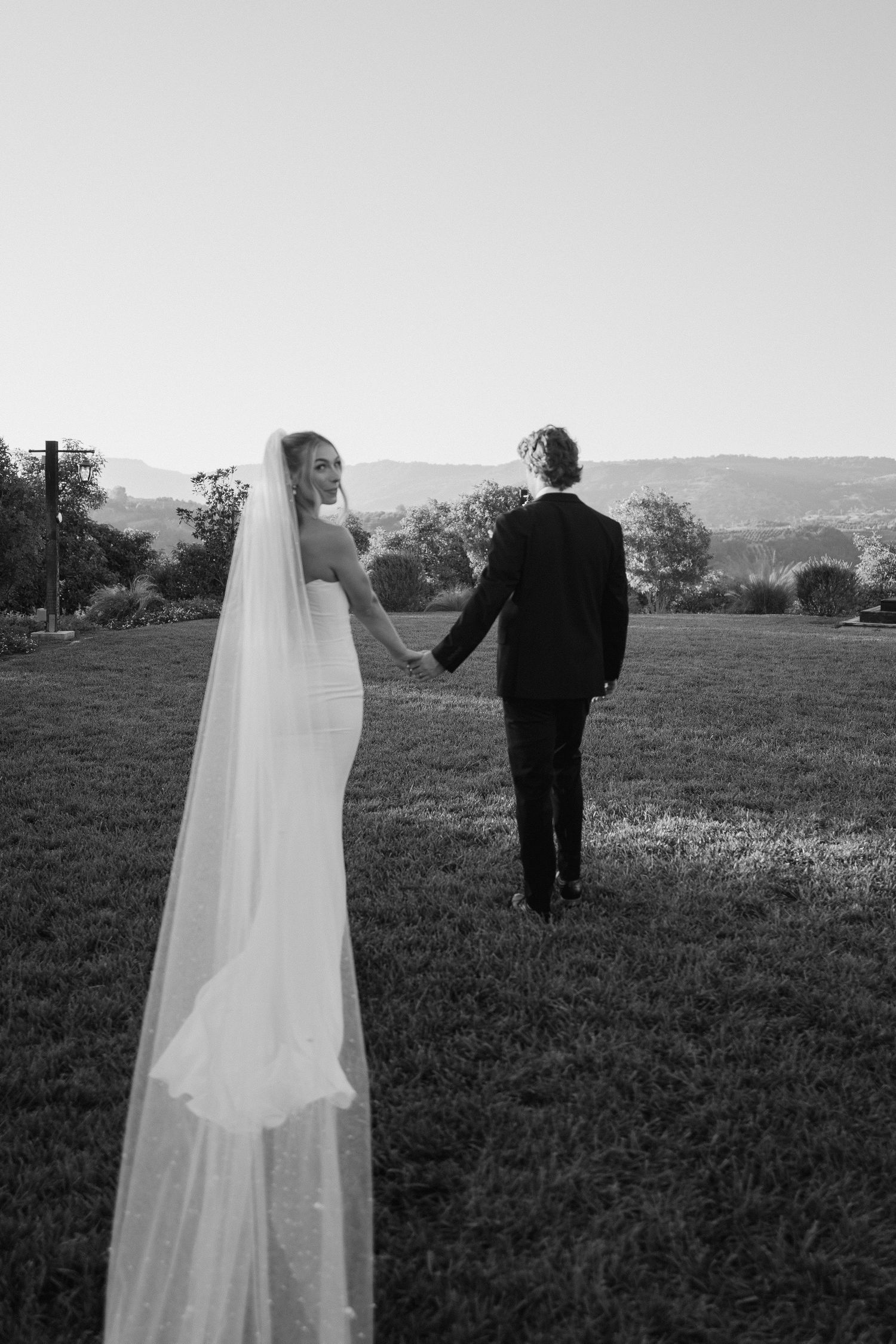 Black and white photo of bride in fitted gown and veil holding hands with groom walking in field.