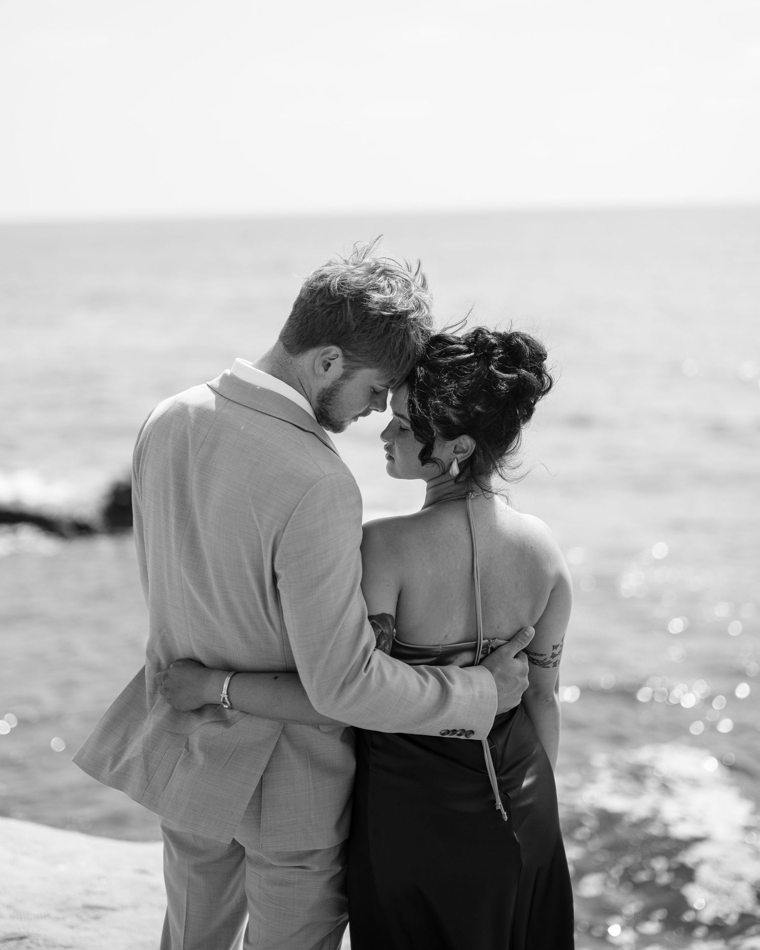 Black and white portrait of a couple embracing intimately by the ocean during a romantic seaside photo session.