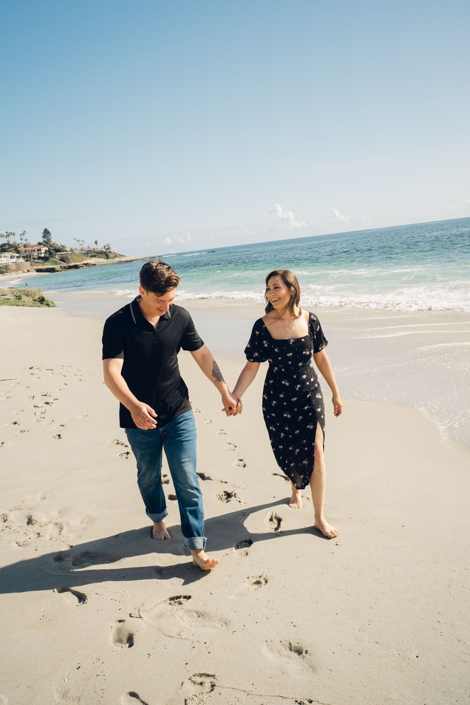 Happy couple holding hands and walking barefoot on sunny sandy beach with blue ocean waves behind them.