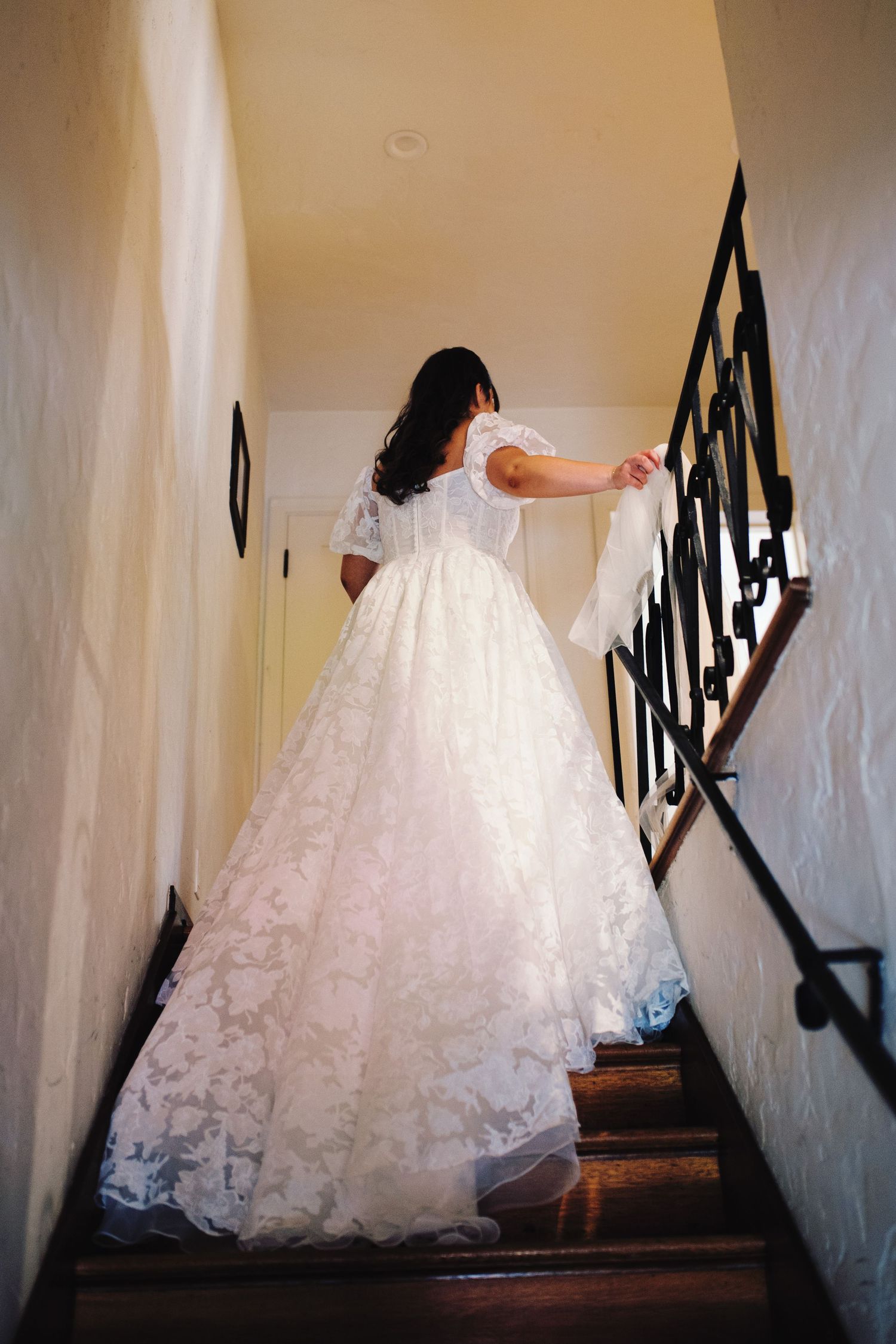 Bride in stunning lace ballgown carries veil while descending staircase with black iron railing.