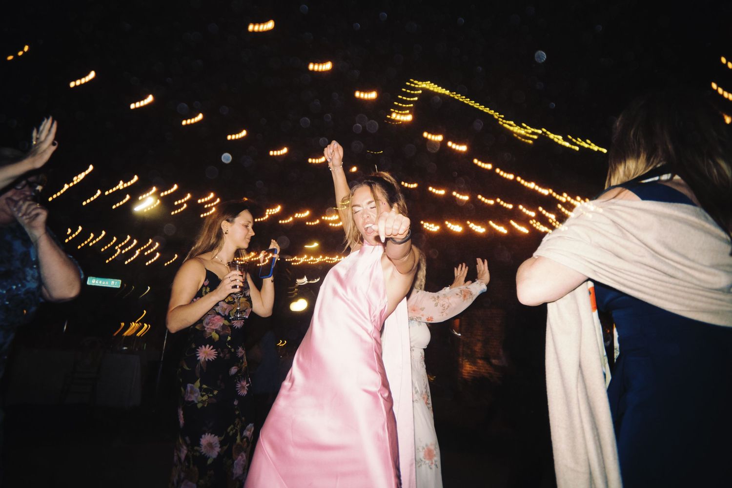 Bride in pink gown dancing joyfully with guests at a nighttime wedding reception.