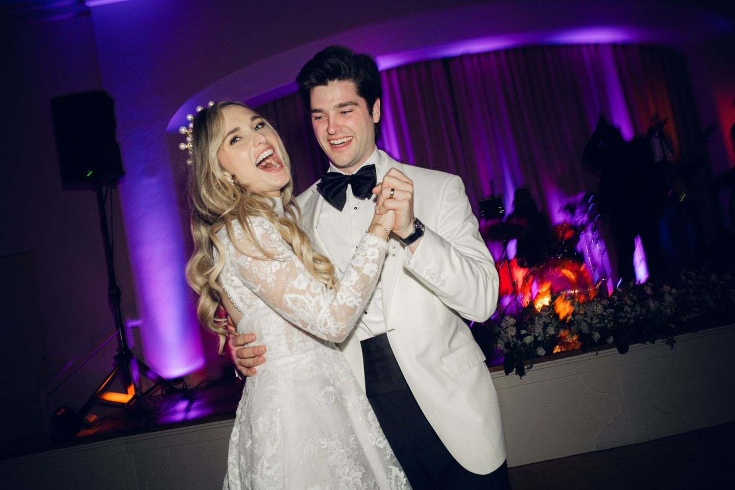 Smiling bride in lace gown and groom in white tuxedo dance together at their wedding reception.