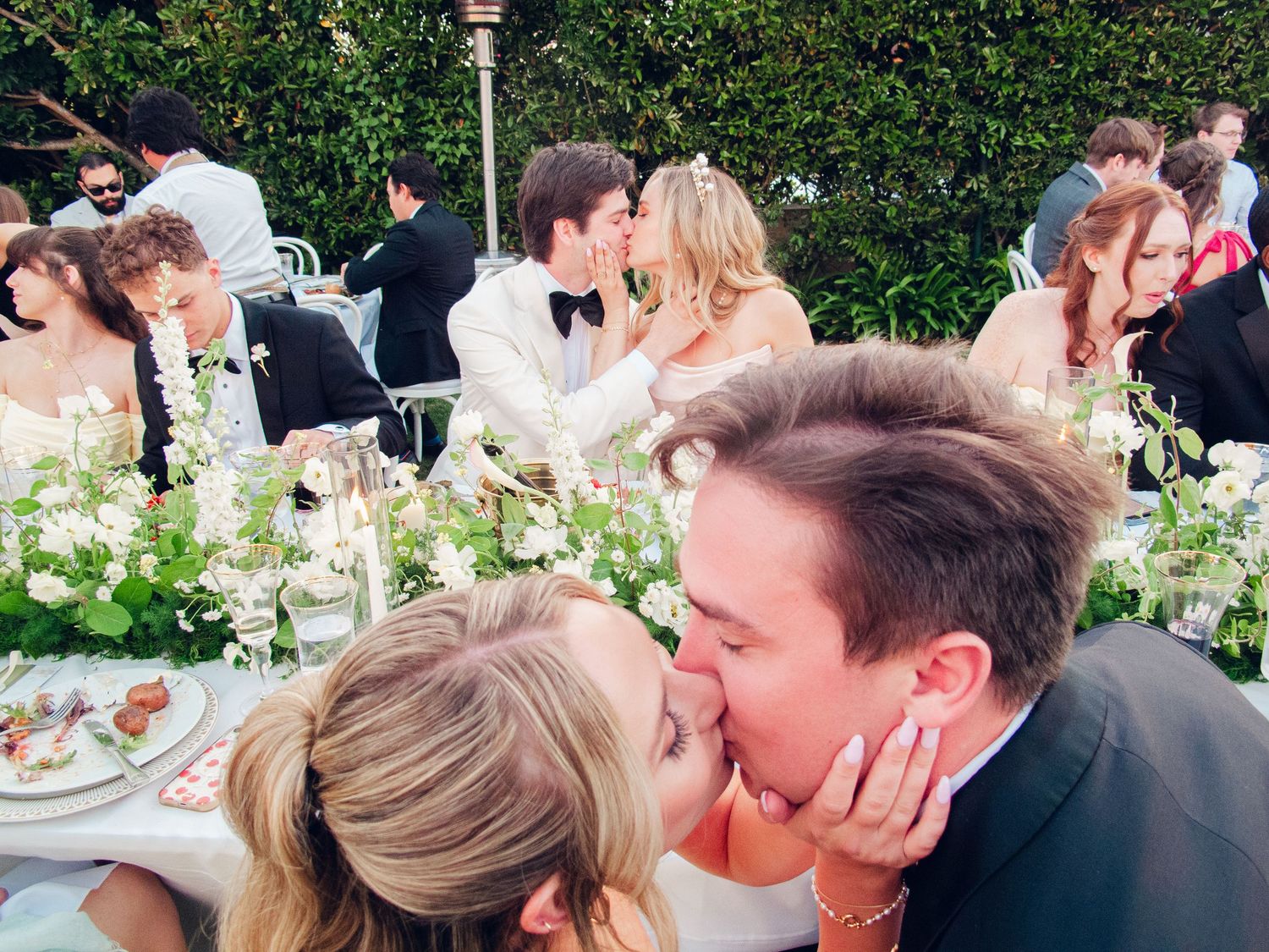 Couple shares a romantic kiss at an outdoor wedding reception with floral centerpieces and guests in background.