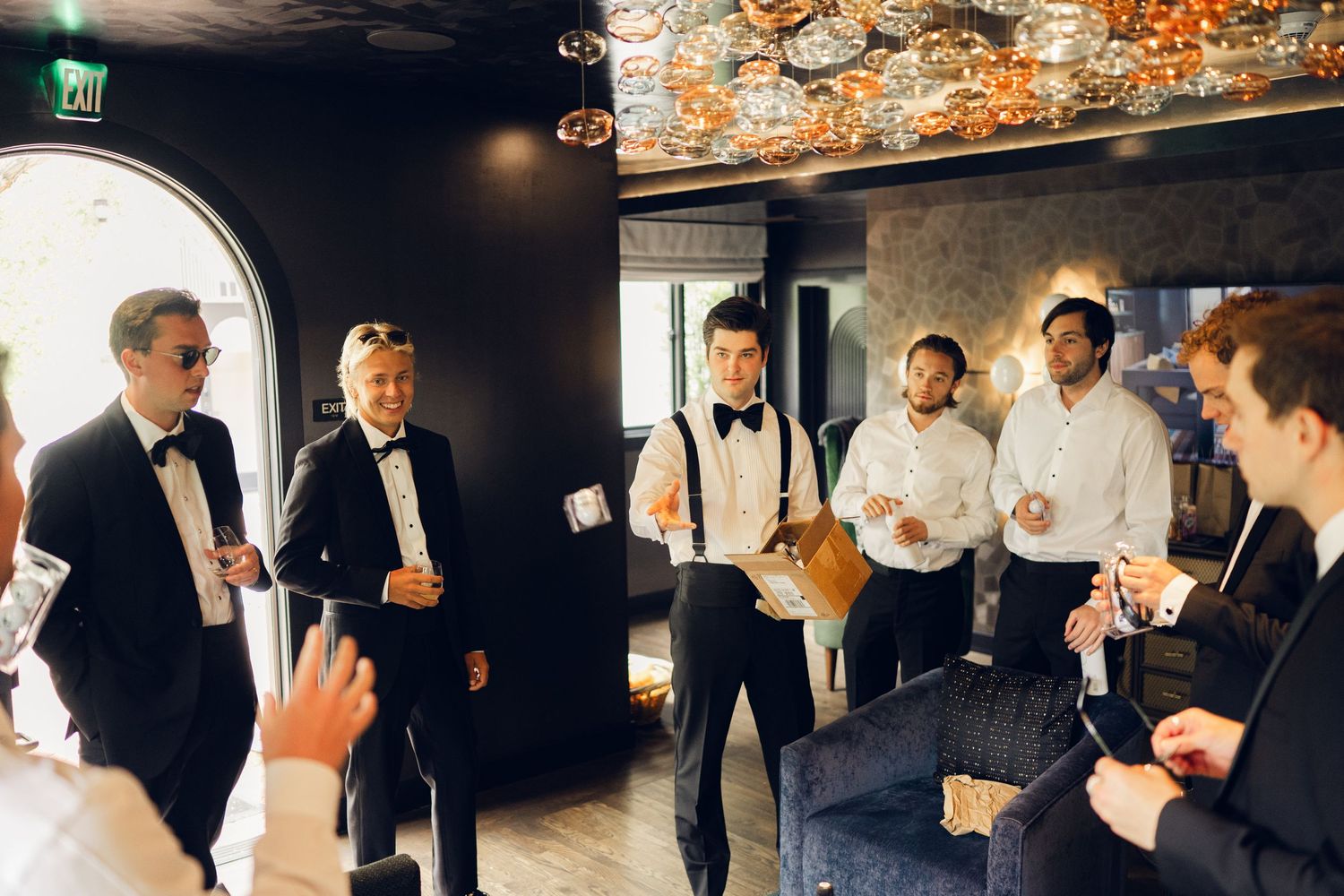 Group of groomsmen in black tuxedos and bow ties socializing inside elegant dark-walled venue before wedding ceremony.