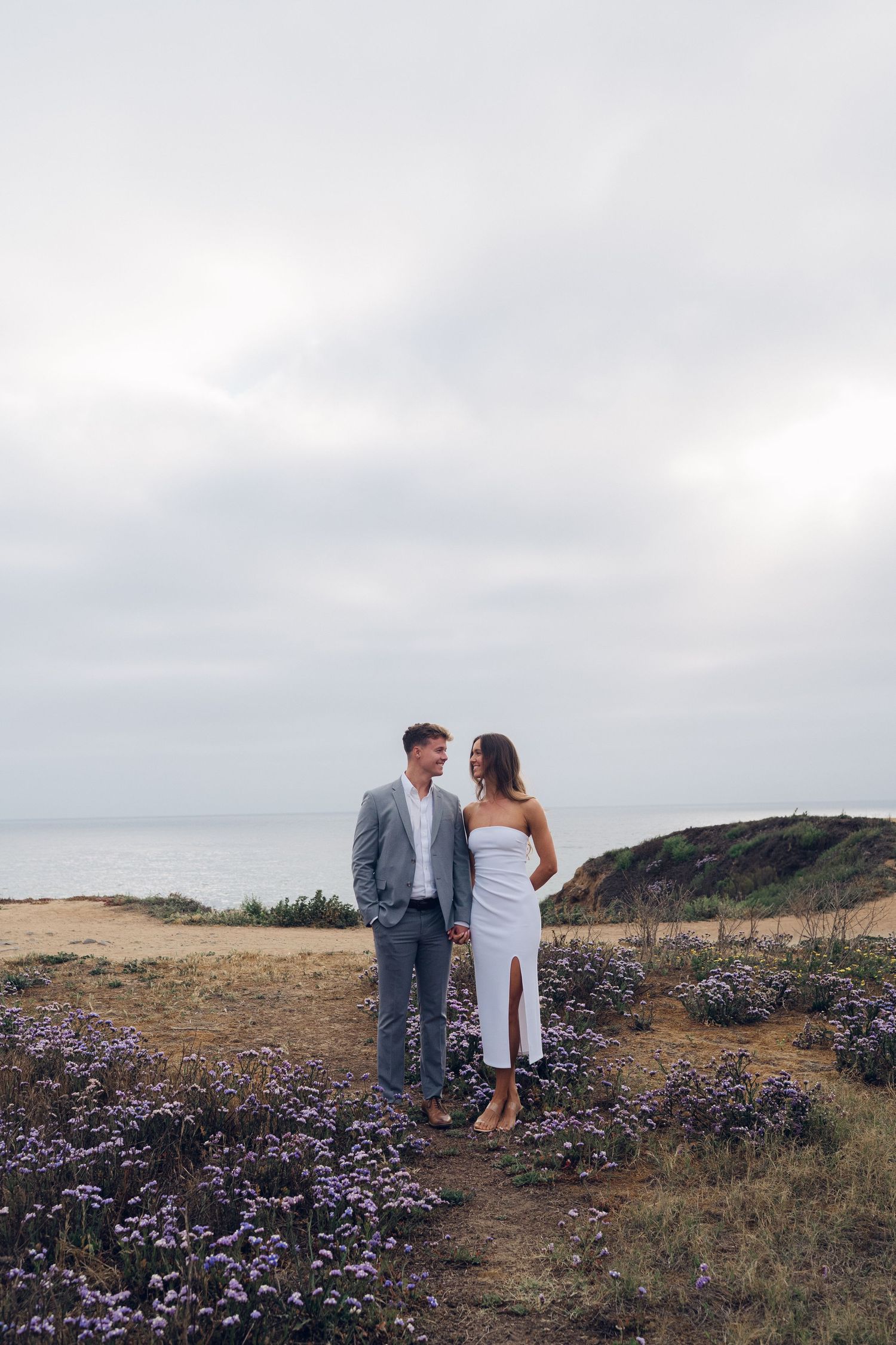 Engaged couple holding hands among purple wildflowers on a coastal cliff with ocean views.