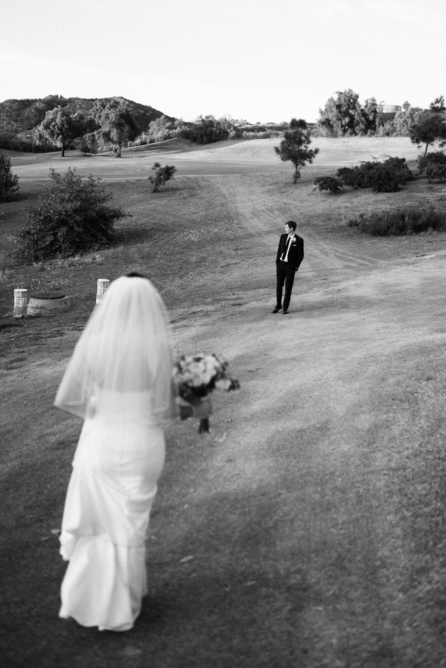 Black and white wedding photo of bride walking toward groom on scenic golf course path, holding bouquet.