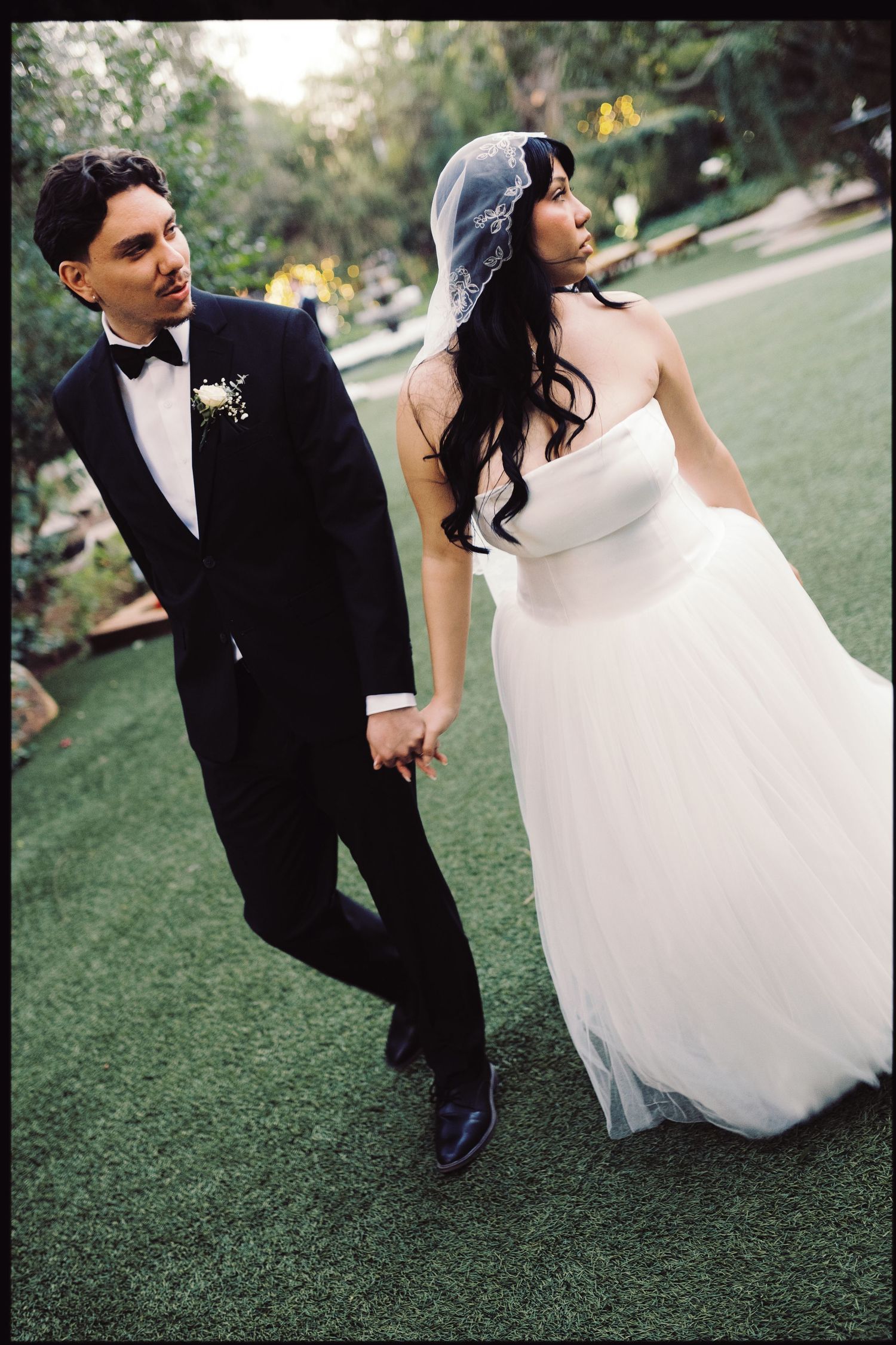 Bride in strapless tulle gown and veiled groom in tuxedo walk together on green lawn.