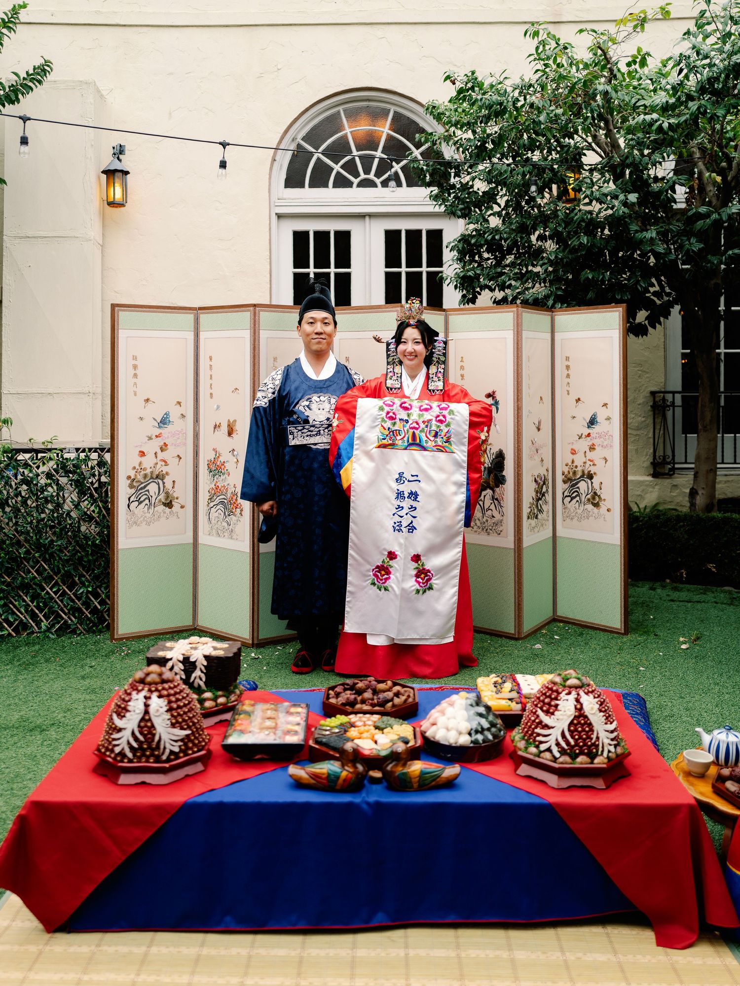 Korean wedding couple in hanbok holding embroidered cloth at traditional paebaek ceremony table.
