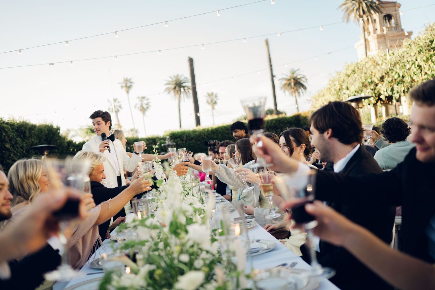 Guests raise glasses in a toast at an elegant outdoor wedding reception with long decorated tables and string lights.