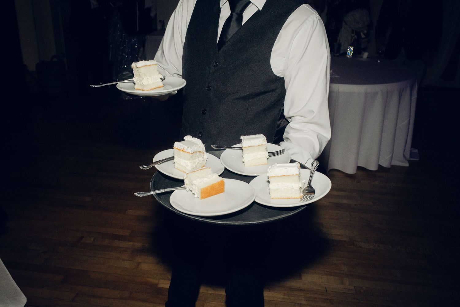 Waiter in formal attire serves multiple plates of white wedding cake slices on a large tray.