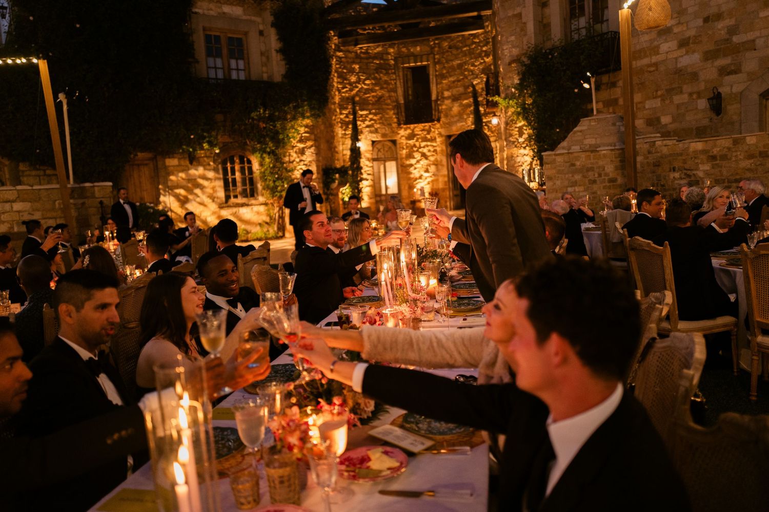 Elegant outdoor wedding reception dinner at night with candlelit tables and guests raising glasses for a toast.