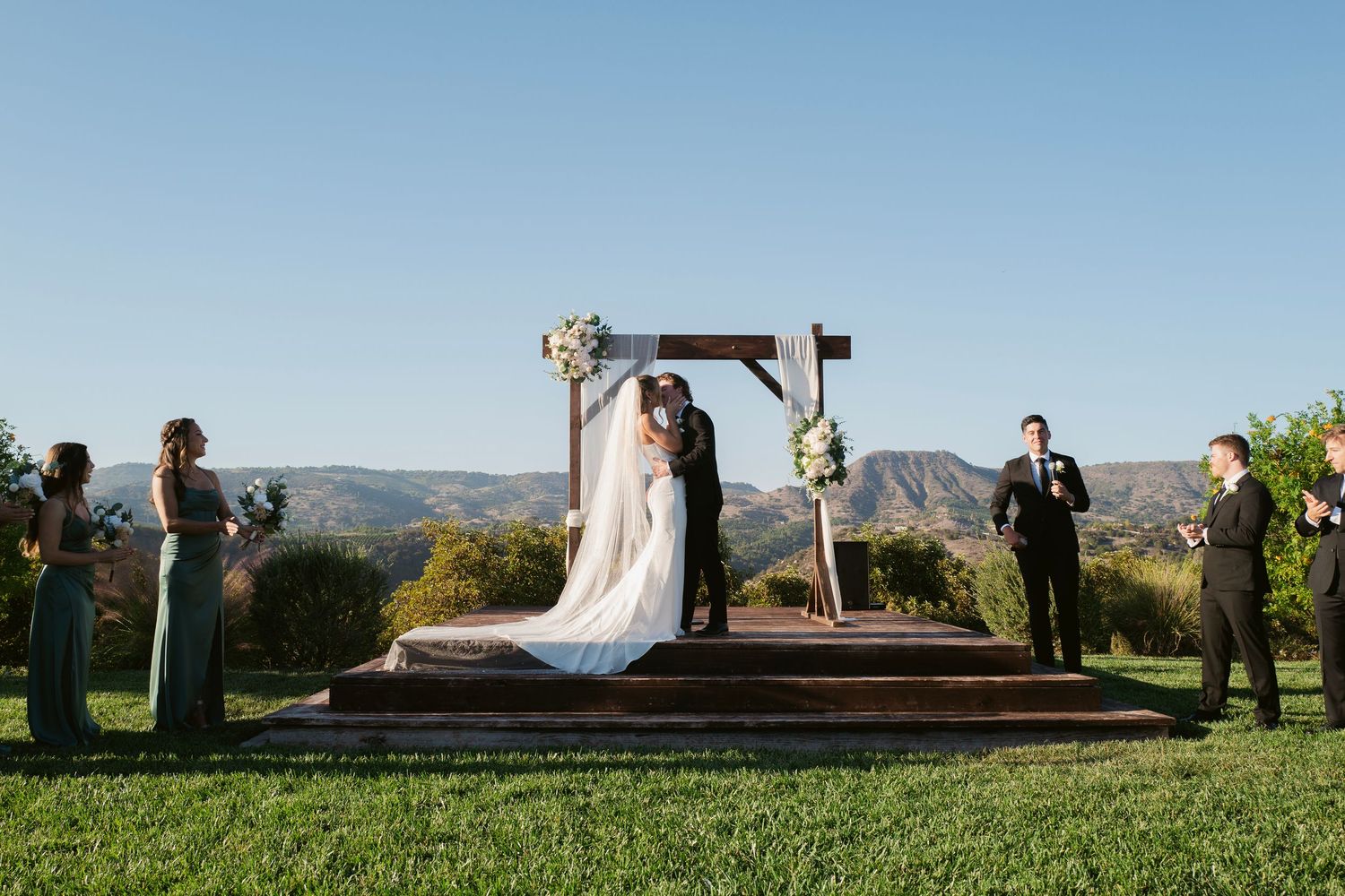 Bride and groom kissing under rustic wooden arch with mountain backdrop at outdoor wedding.