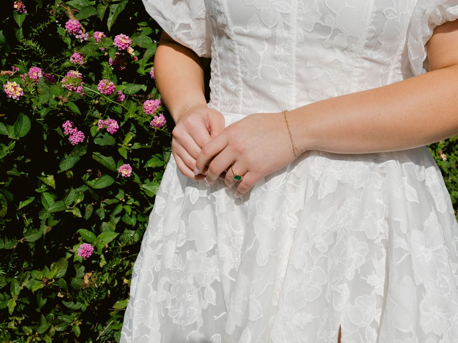 Close-up of bride in white lace wedding dress with delicate bracelet, hands clasped near pink garden flowers.