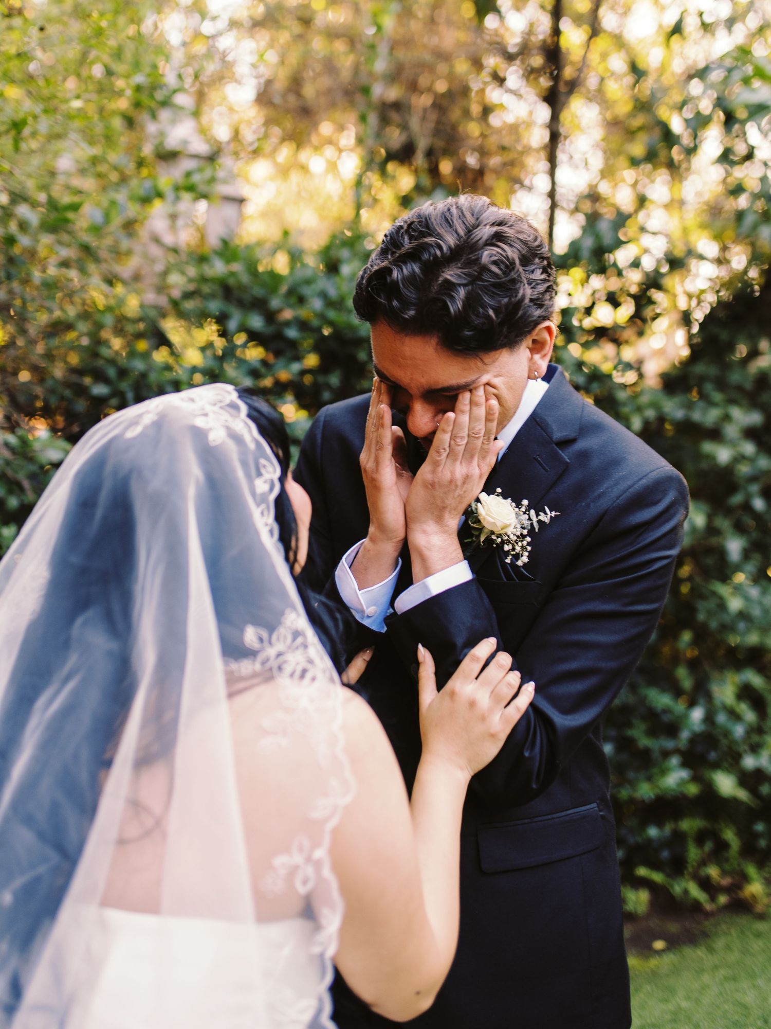 Tearful groom covering face with hands during emotional first look with veiled bride.
