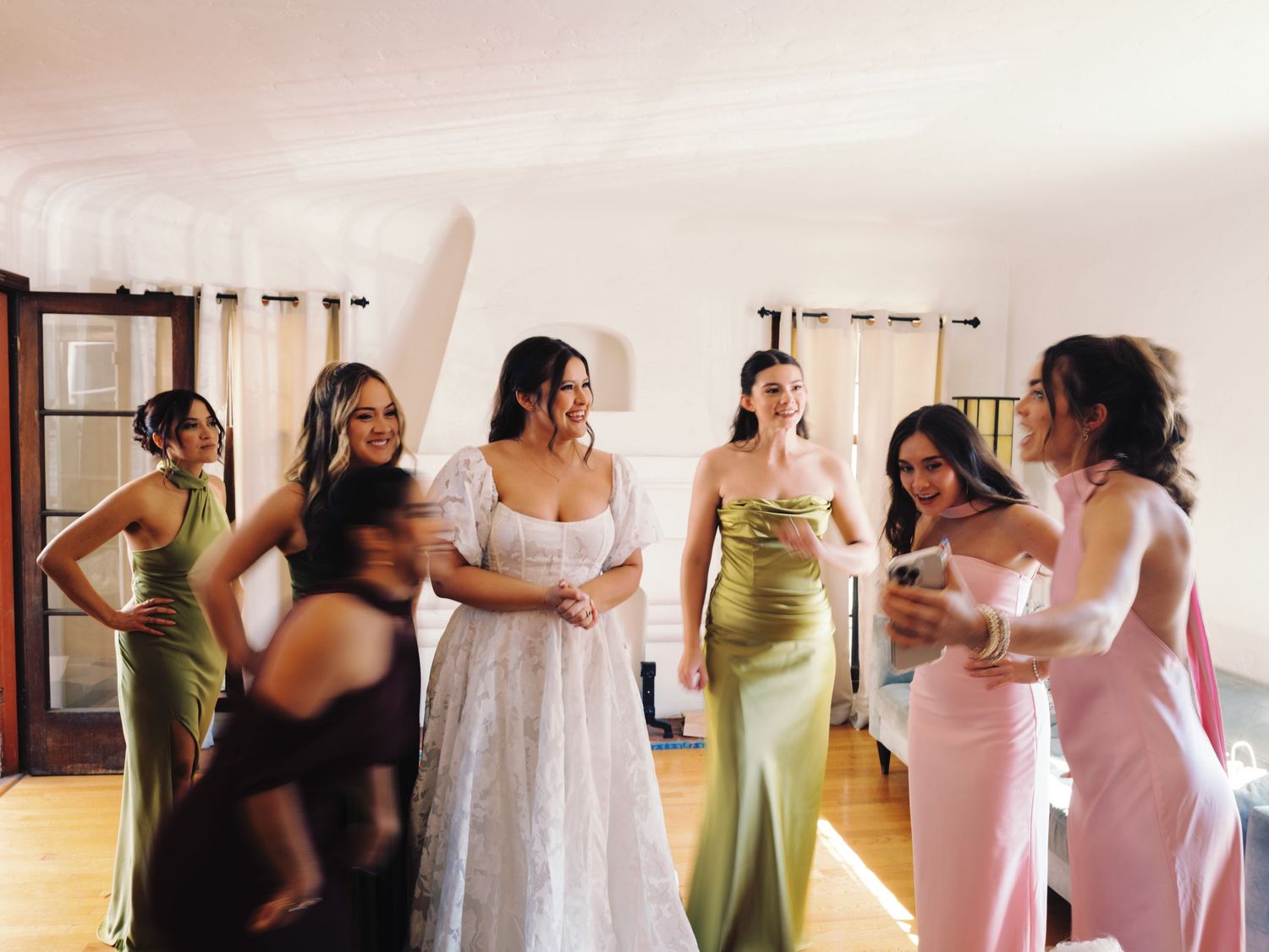 Bride in white lace gown surrounded by bridesmaids in colorful satin dresses during wedding preparations indoors.
