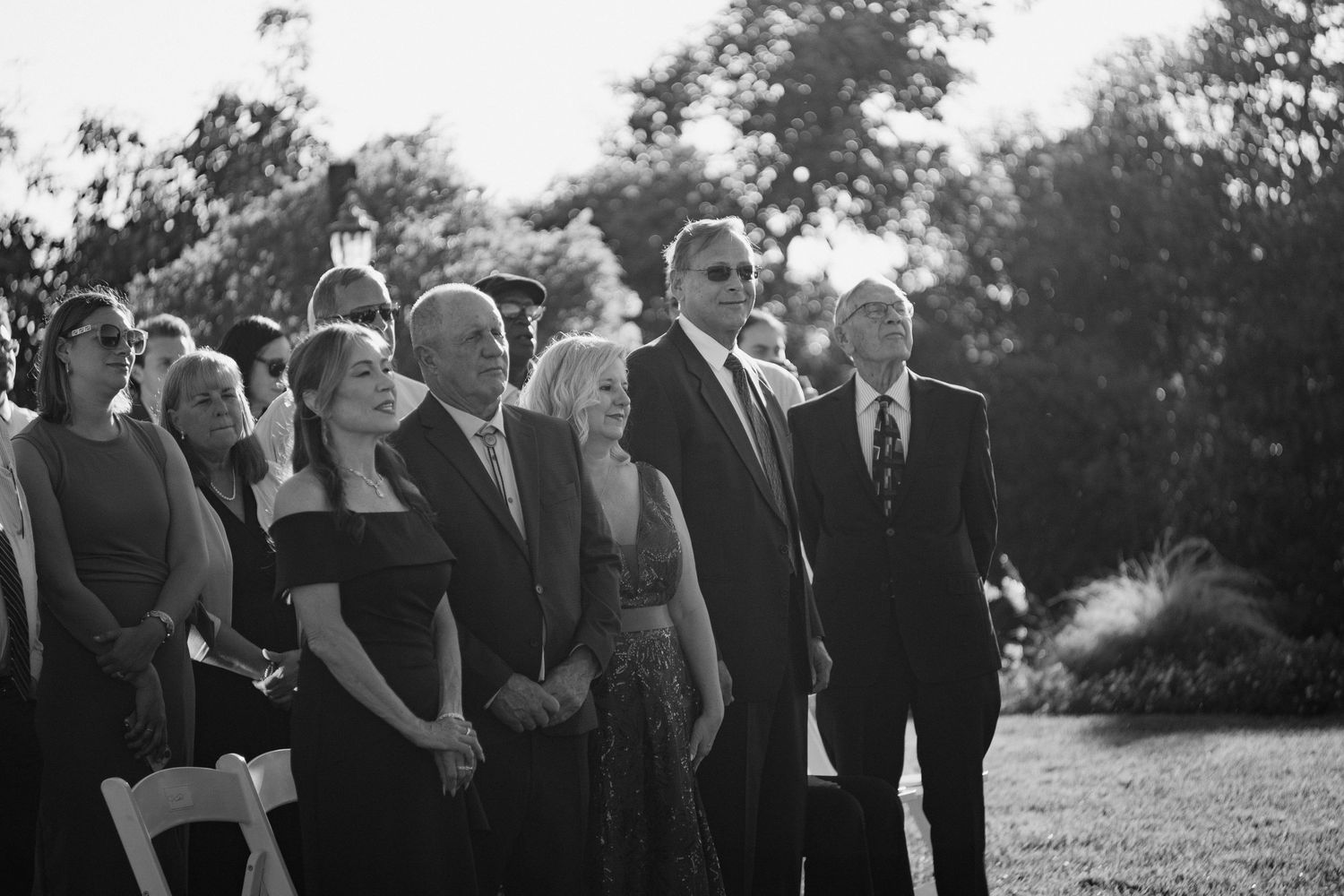 Crowd of elegantly dressed wedding guests standing during an outdoor ceremony in black and white.