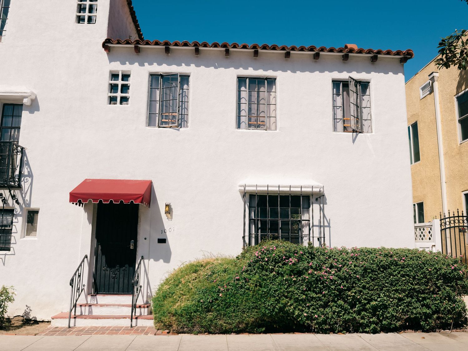 White Spanish Colonial Revival style home with red awning, iron railings, and lush green hedges in Los Angeles.