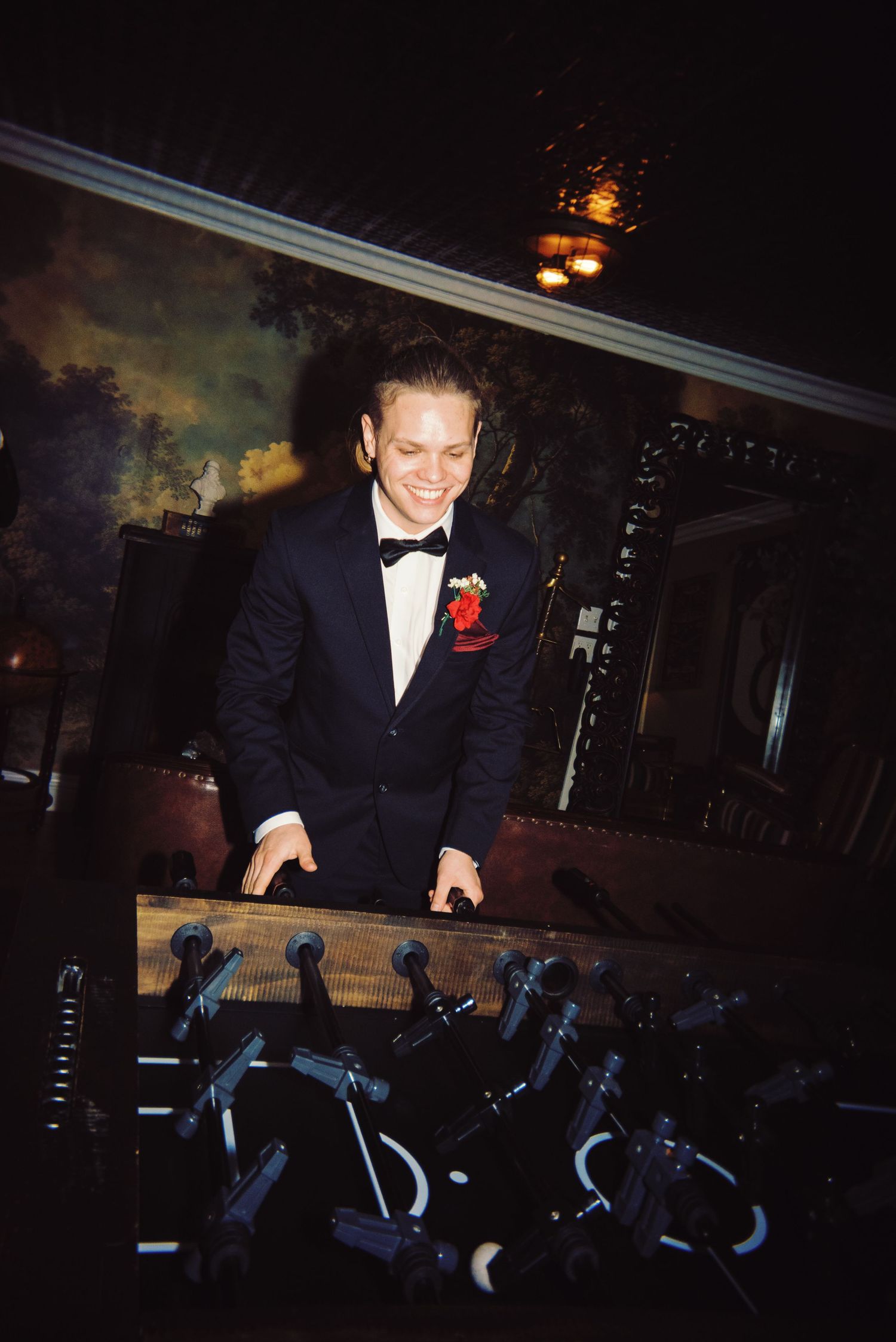 Smiling groom in tuxedo with red boutonniere playing foosball at wedding reception.