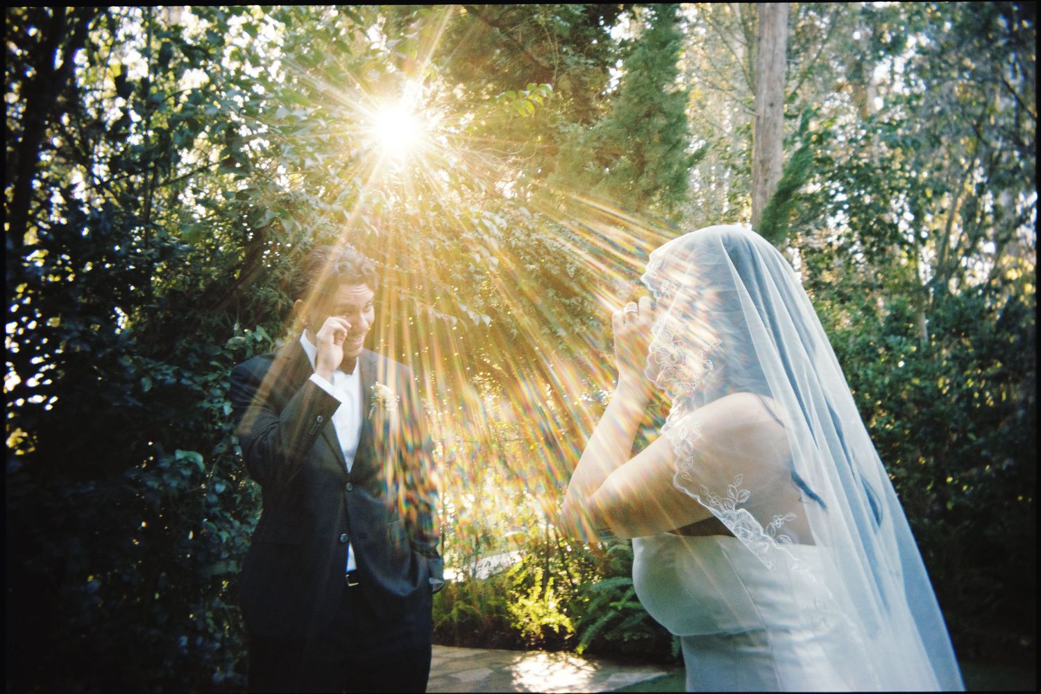 Bride and groom emotional first look with stunning sunburst through garden trees.