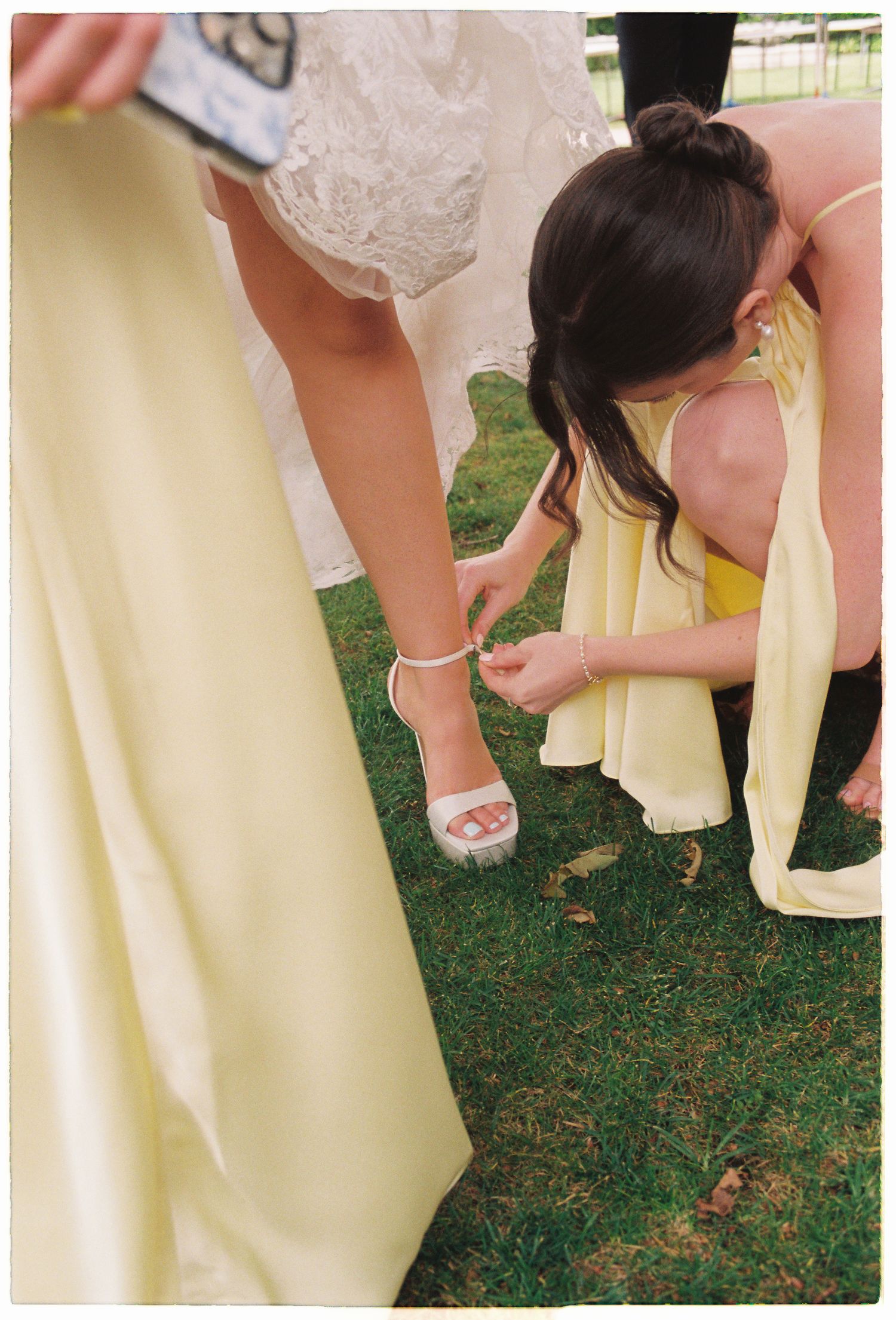 Bridesmaid in yellow dress kneels on grass to fasten the strap on the bride's white wedding heel shoe.