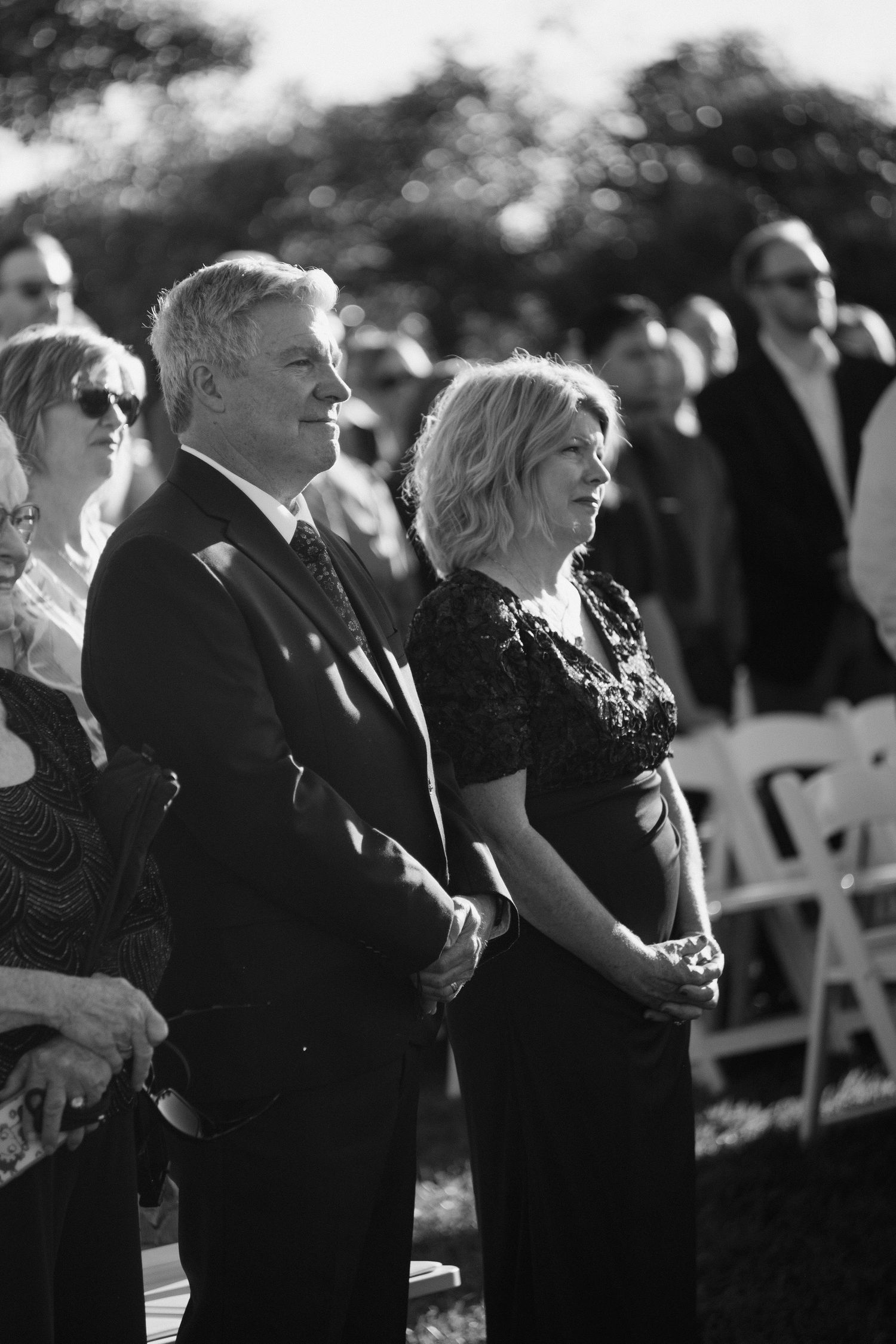 Emotional wedding guests in formal attire watching an outdoor ceremony in black and white photography.