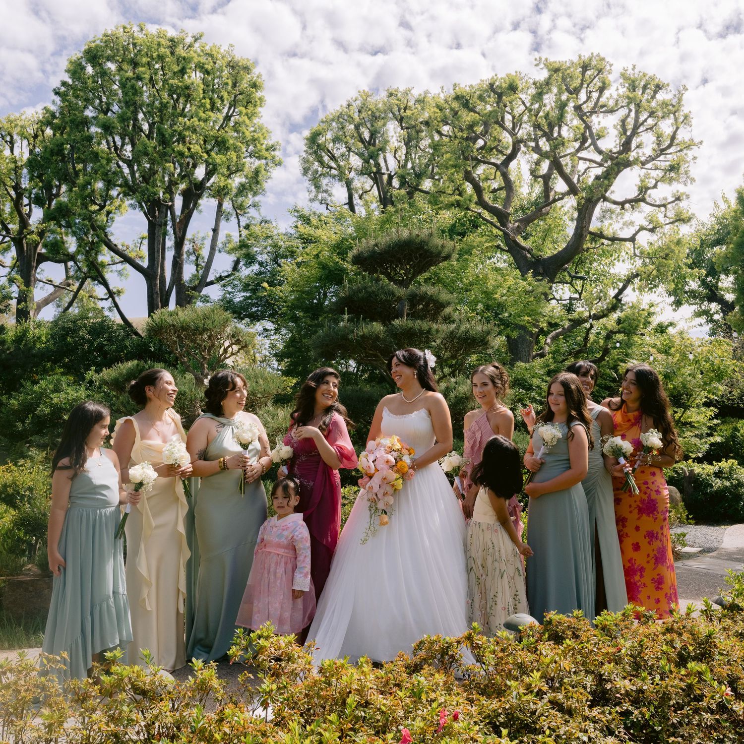 Bride in white gown poses with bridesmaids in mismatched pastel dresses in a lush garden setting.