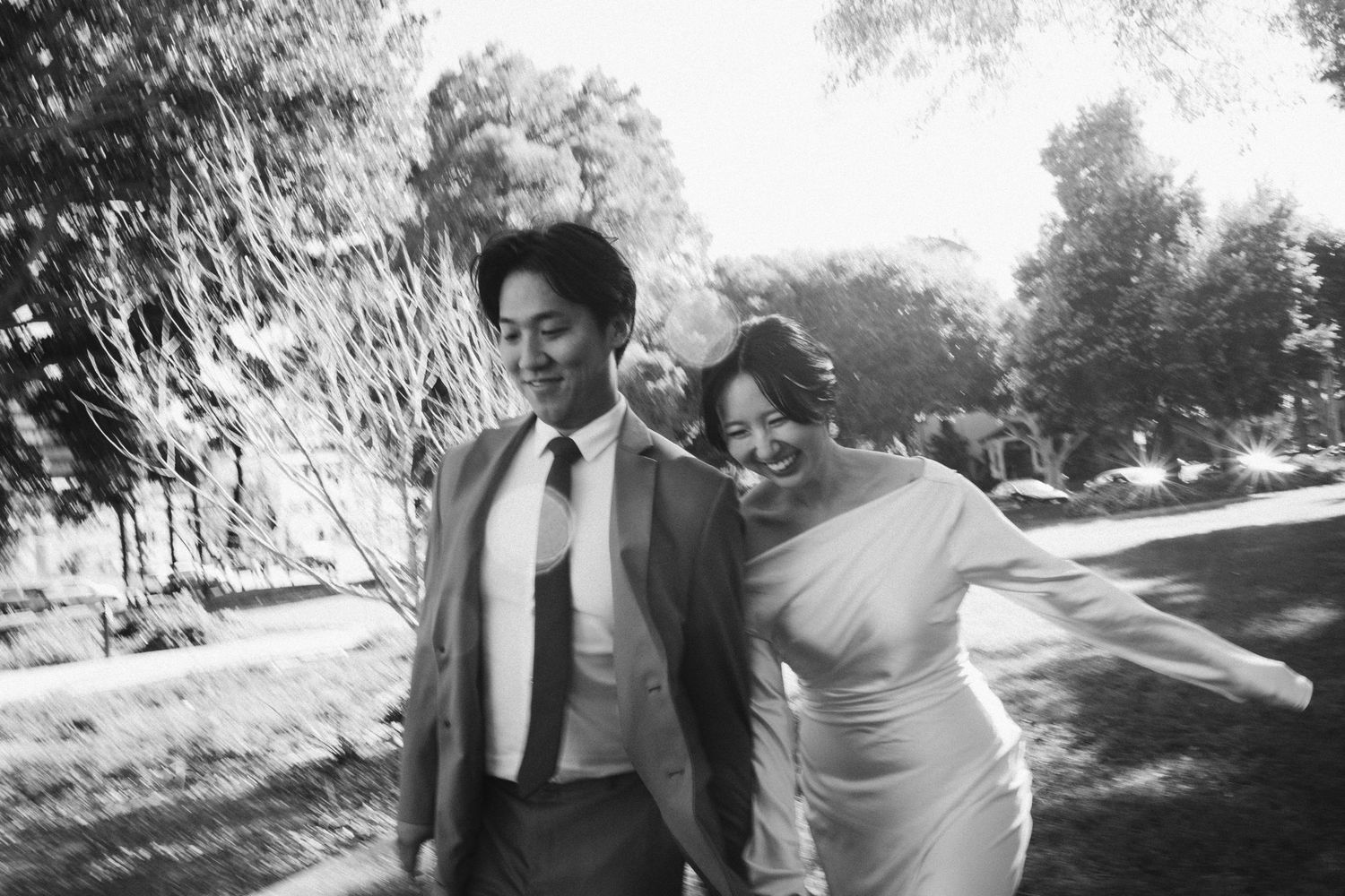Black and white photo of a smiling couple in wedding attire walking outdoors among trees on their wedding day.