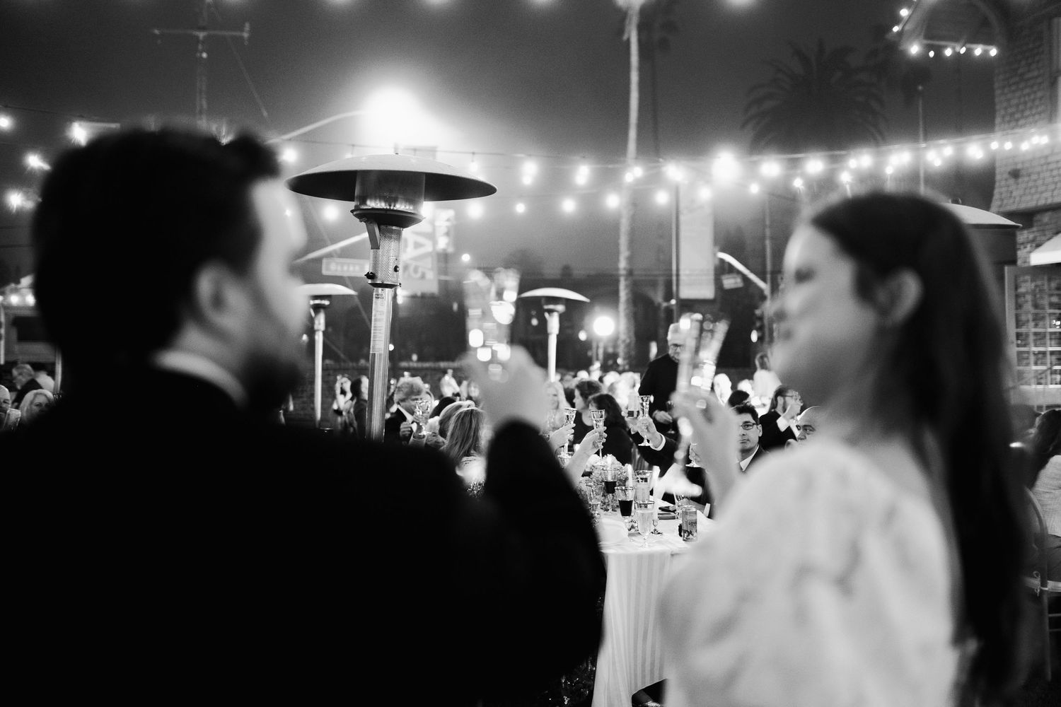 Black and white photo of wedding guests seated at outdoor reception tables under warm string lights at night.