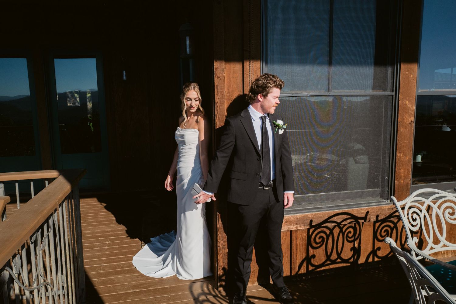 Bride and groom hold hands on sunlit balcony with mountain views on wedding day.