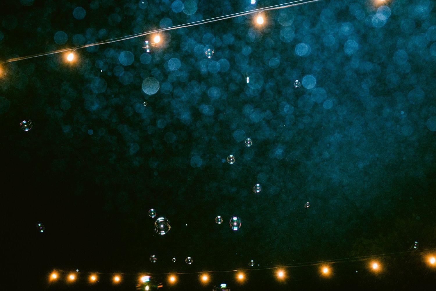 Magical bokeh shot of bubbles floating under string lights at outdoor wedding reception at night.